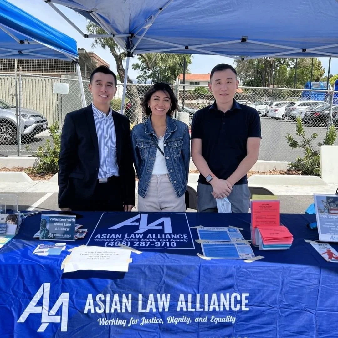Three people standing behind a table with Asian Law Alliance banners at an outdoor event under a blue tent. The table has brochures, flyers, and a sign. The background shows a parking lot with cars and a chain-link fence.
