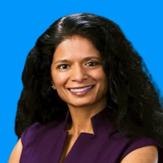 Close-up portrait of a woman with long, curly black hair, smiling, wearing a purple sleeveless top, against a blue background.
