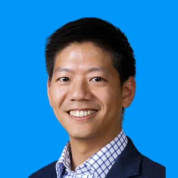 Headshot of a smiling Asian man in a business suit and checkered dress shirt against a blue background.