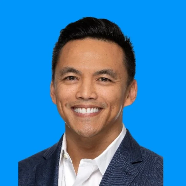 A smiling man with short dark hair dressed in a white shirt and dark blazer against a blue background.