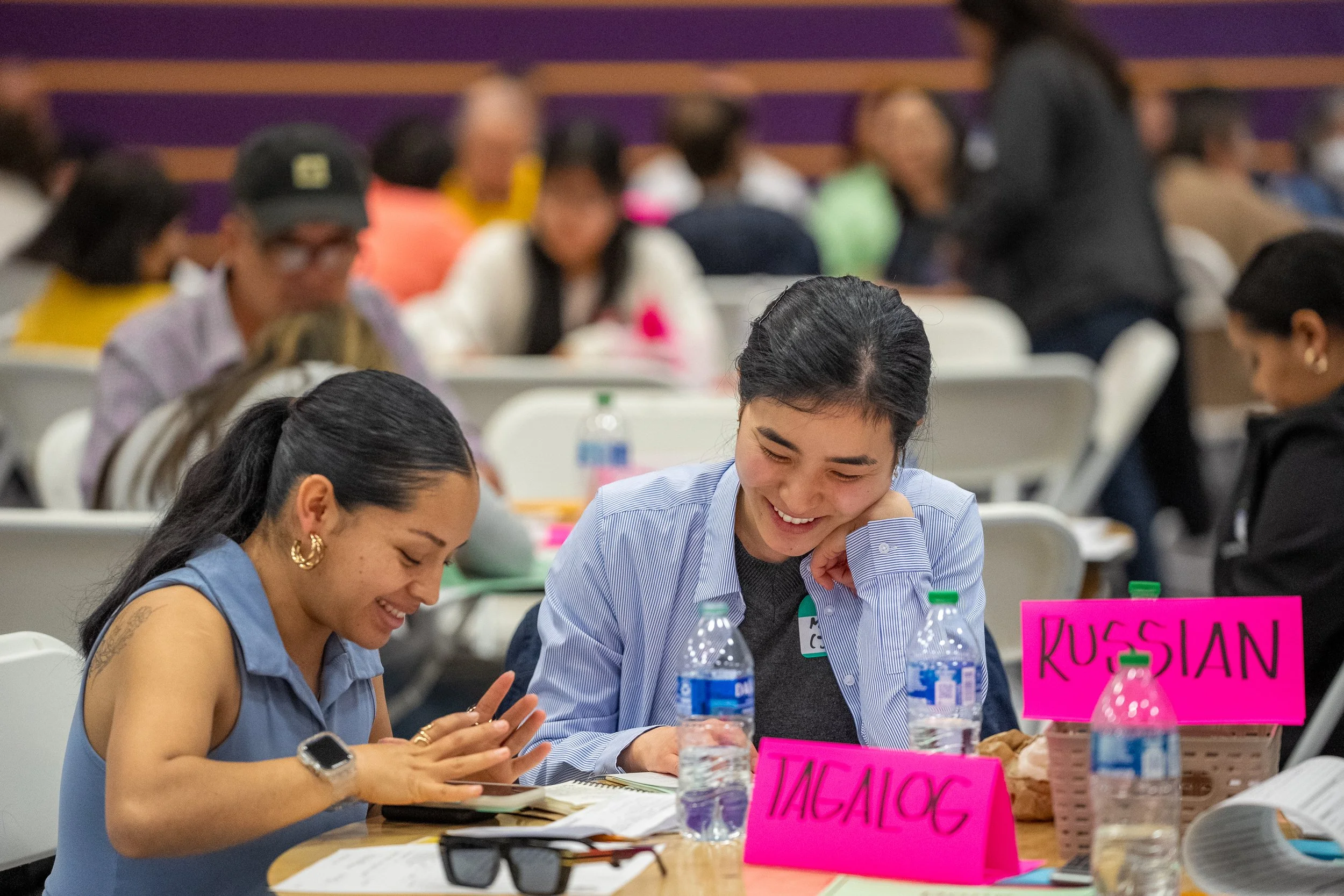 Two women smiling and looking at a phone at a table with water bottles, papers, and pink name tags labeled 'JACKSON' and 'KUSHAN' during an event.