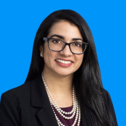 A woman with long black hair, glasses, and pearl necklace smiling against a blue background.