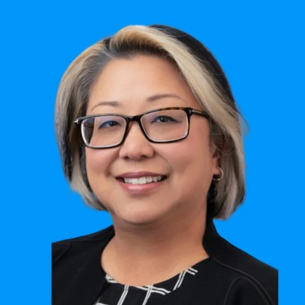 Portrait of a woman with glasses, blonde and black hair, smiling, wearing a black jacket and patterned top, against a blue background.