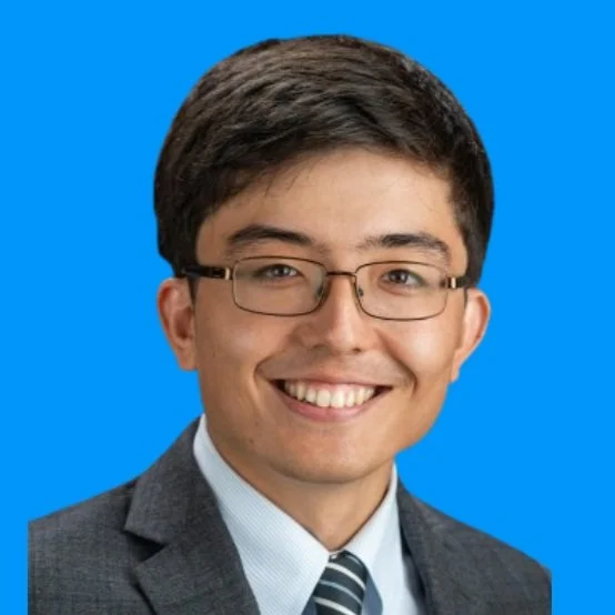 A young man with dark hair, glasses, and a suit smiling against a blue background.