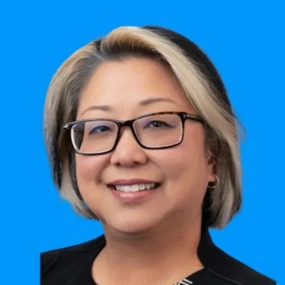 Portrait of a woman with glasses, blonde and black hair, smiling, wearing a black jacket and patterned top, against a blue background.