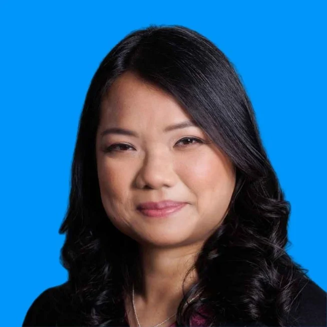 Close-up portrait of an Asian woman with long black hair, smiling softly against a solid blue background.
