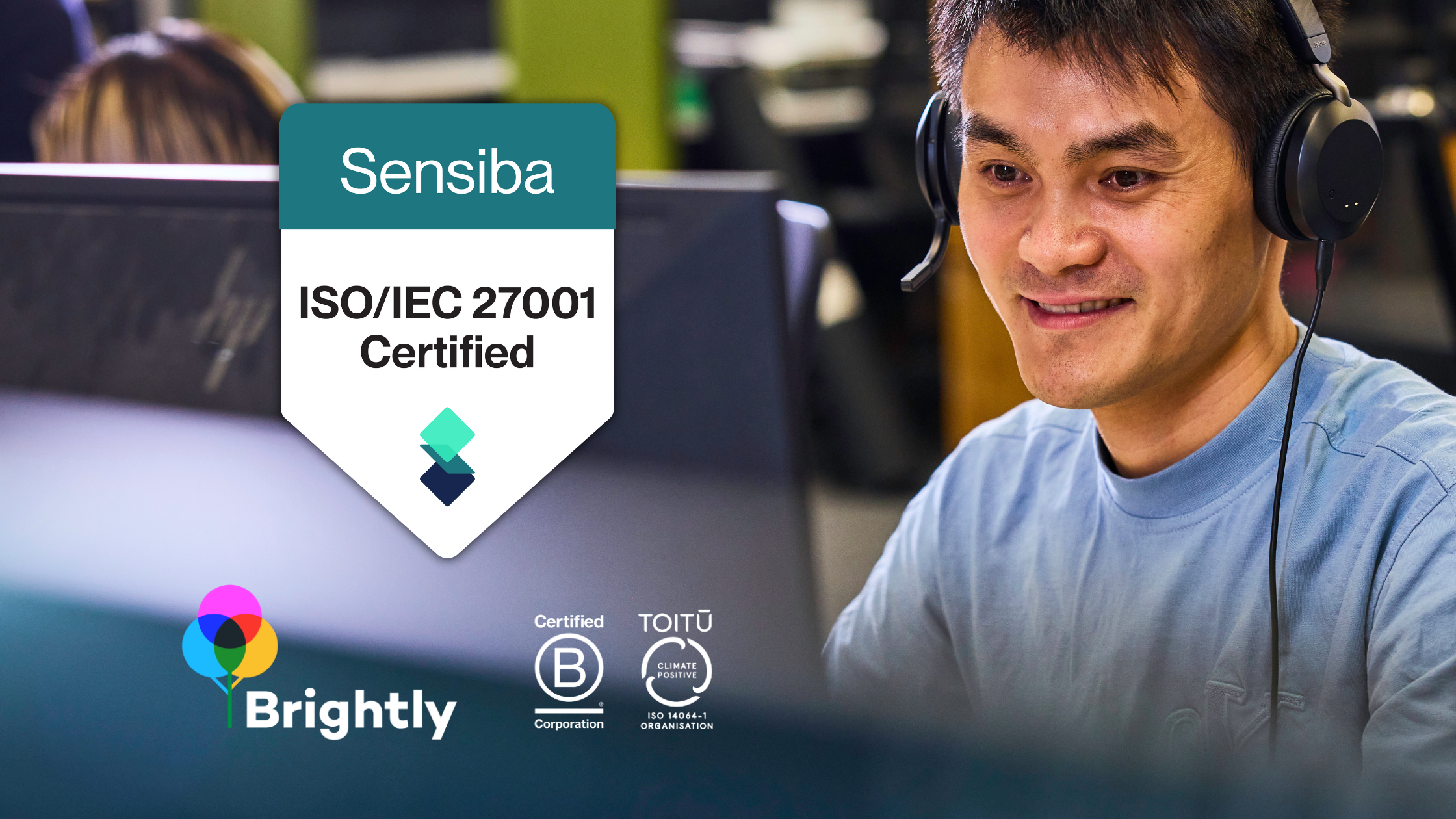 A man wearing a headset working at a computer with certification logos for Brightly, ISO 27001:2022, and climate positive organisation on the screen.