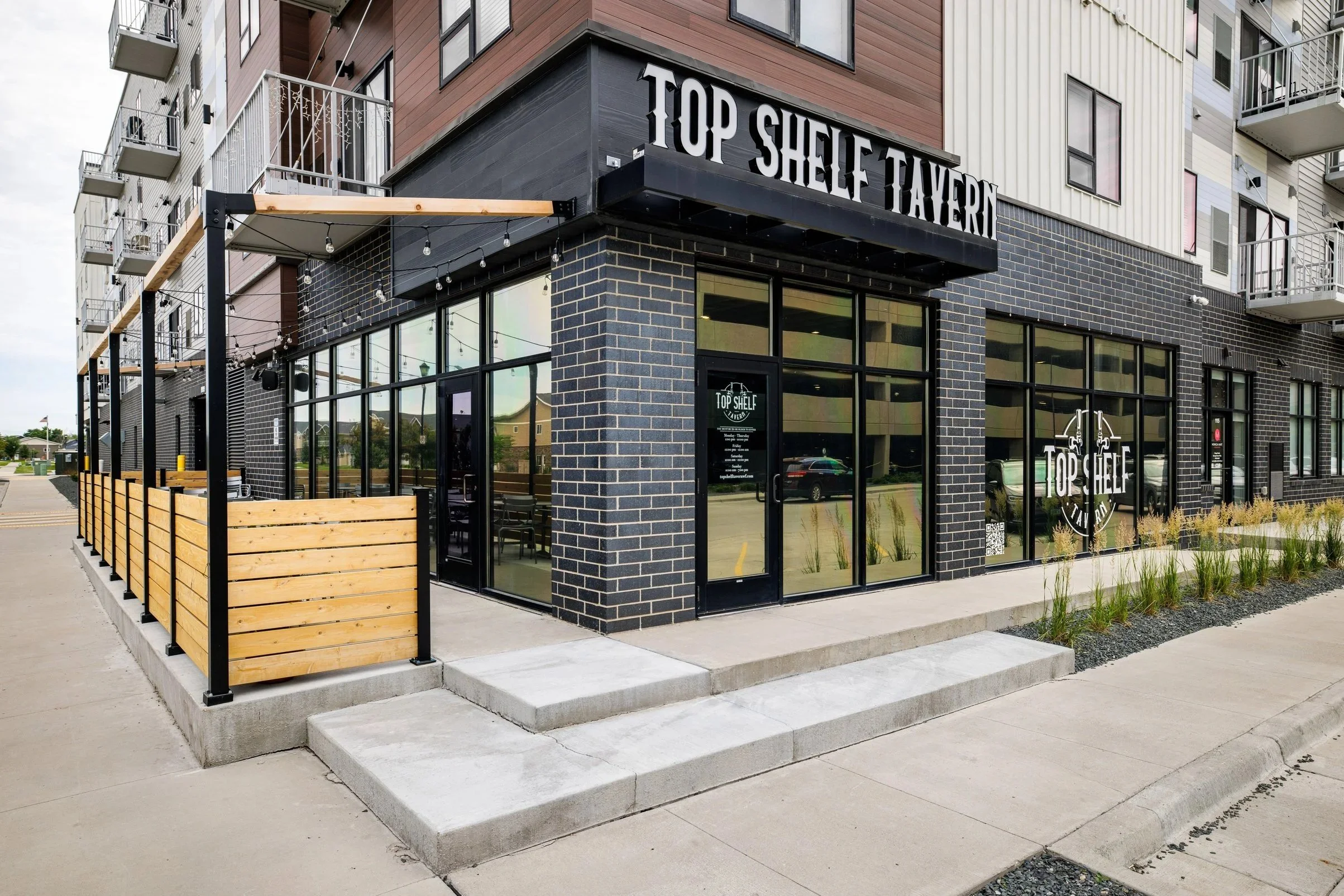 Outside view of "Top Shelf Tavern" with outdoor seating, modern building facade, and large glass windows.
