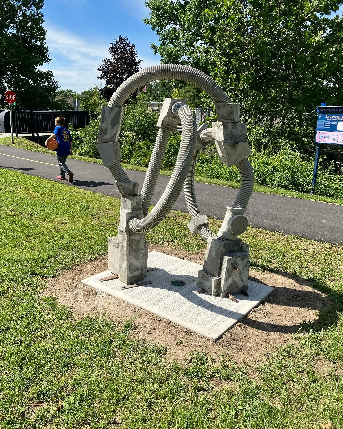 Abstract metal sculpture with flexible ducts on a concrete base near a pathway, with a person carrying a basketball walking towards a stop sign, surrounded by trees and grass.