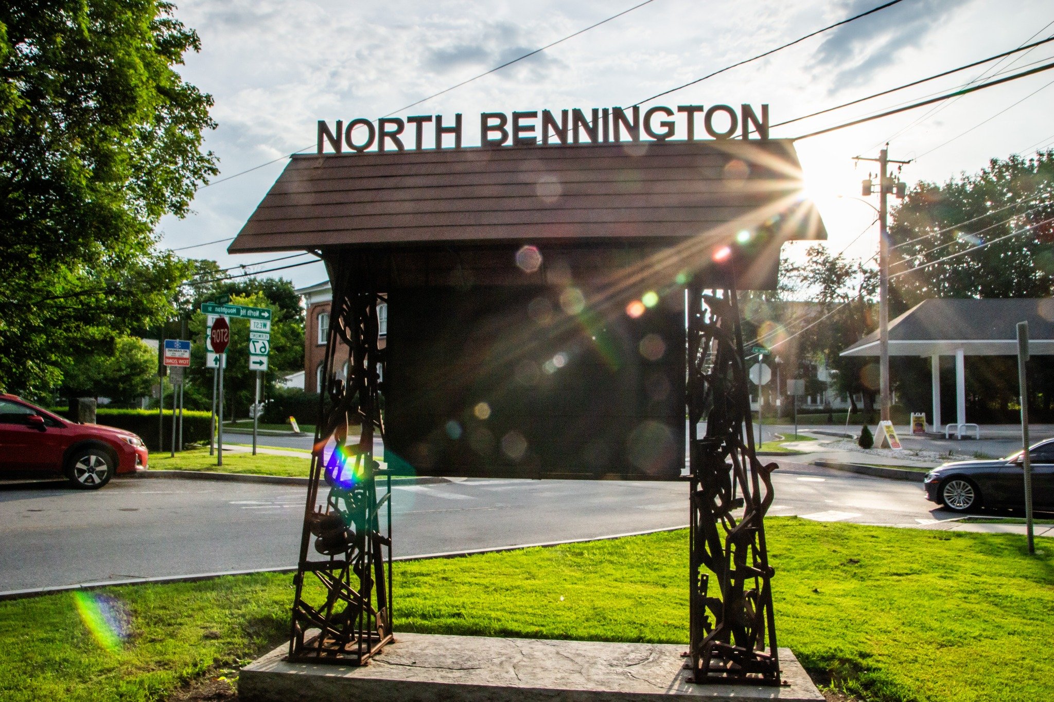 Outdoor event with people walking on grass and a dirt path among trees, tents, and colorful sculptures, including a large green bottle and a metal structure.
