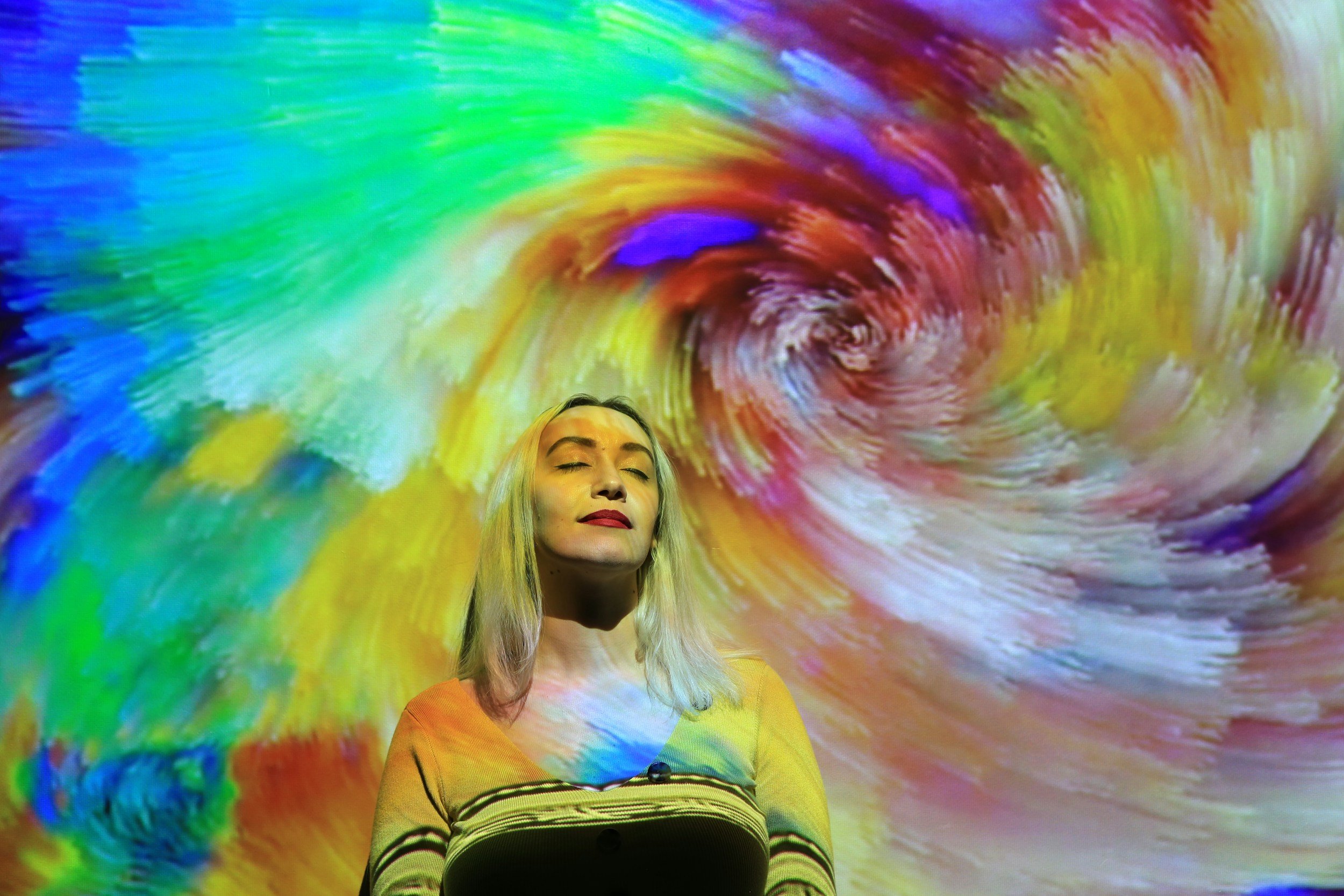 A woman with long blonde hair and closed eyes standing in front of a colorful, swirling psychedelic background.