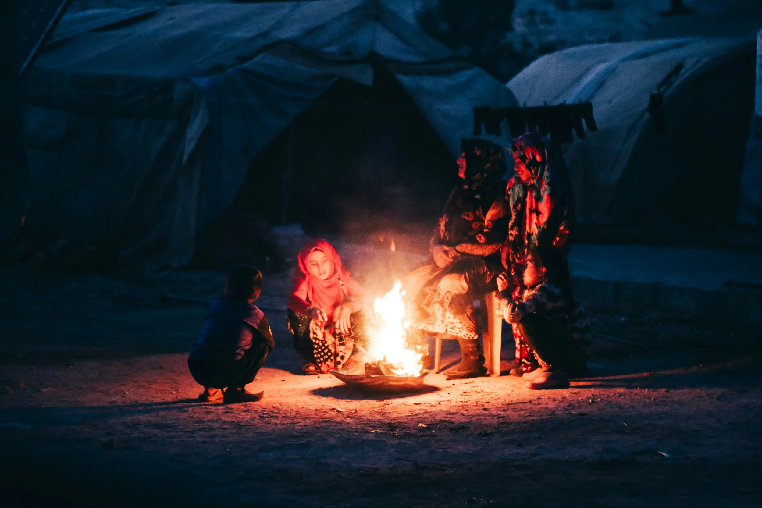 Children gathered around a small campfire outdoors at night, surrounded by snow, with a tent in the background.