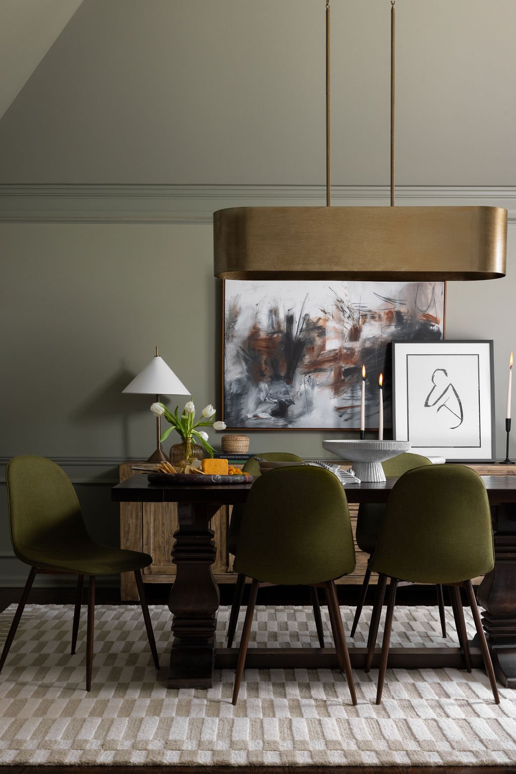Dining room with a dark wood table surrounded by six green upholstered chairs, a large white and gray abstract painting, a framed minimalist line art portrait, candles, a table lamp, a vase with white flowers, and a bowl with fruit on the table. Overhead, a large wooden pendant light hangs.