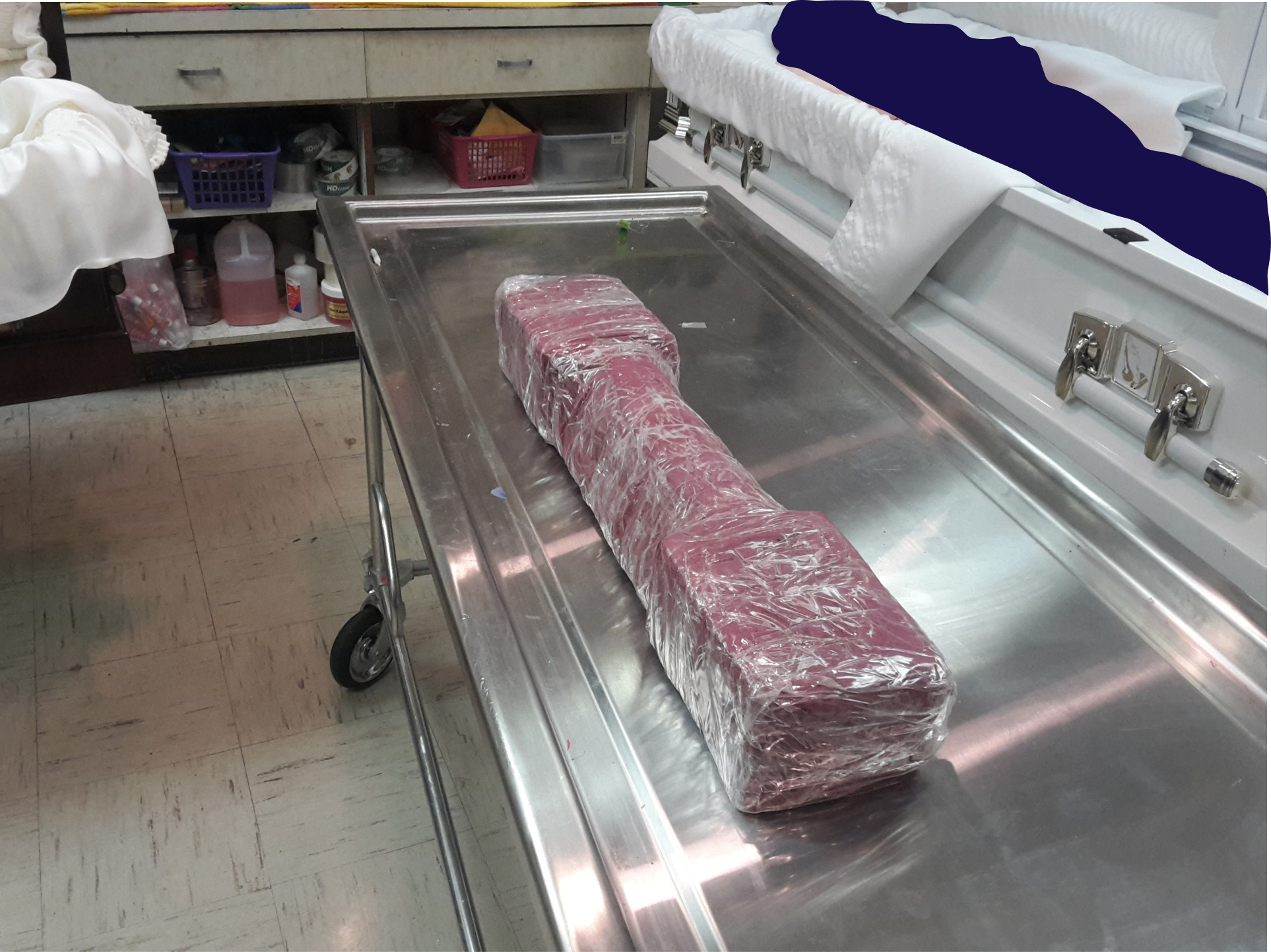 A large piece of meat wrapped in plastic on a stainless steel hospital gurney in a sterile environment