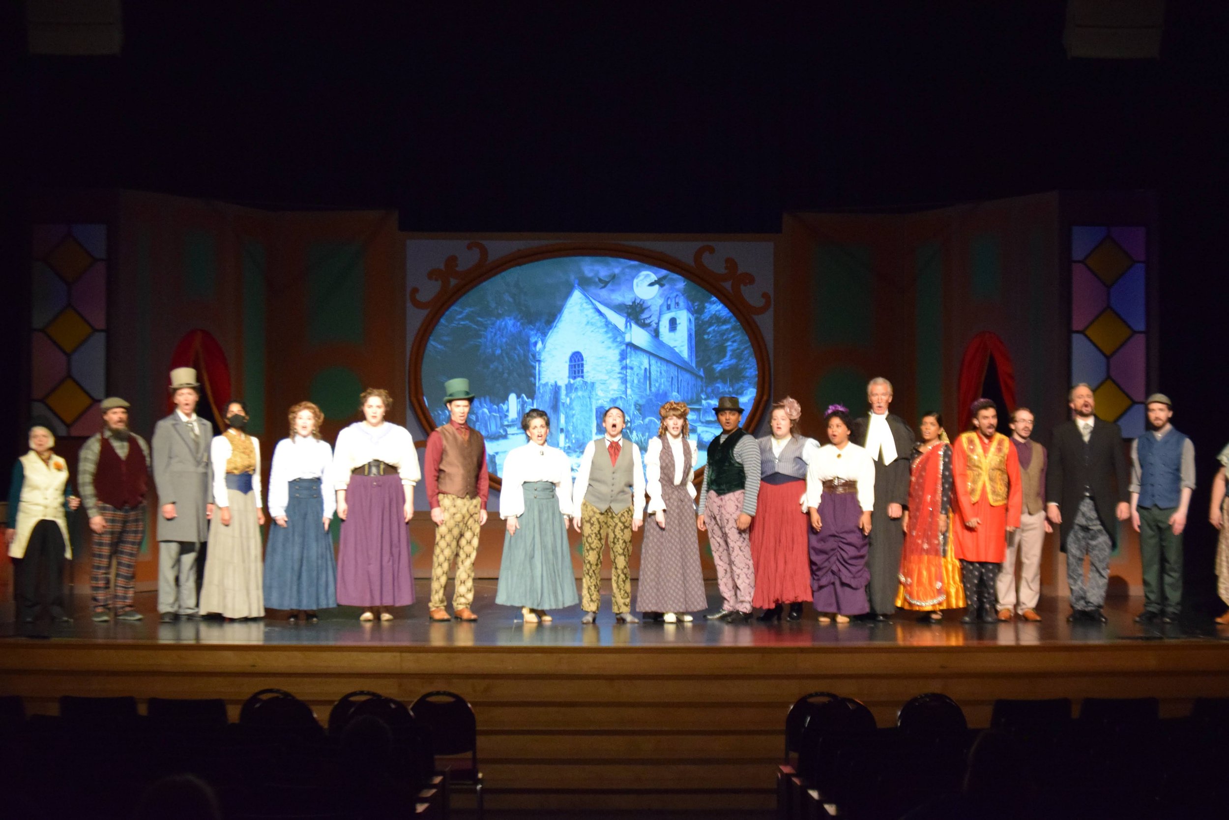 The cast of The Mystery of Edwin Drood stand in a line on the lip of the stage before an image of a tomb is projected on the screen behind them.