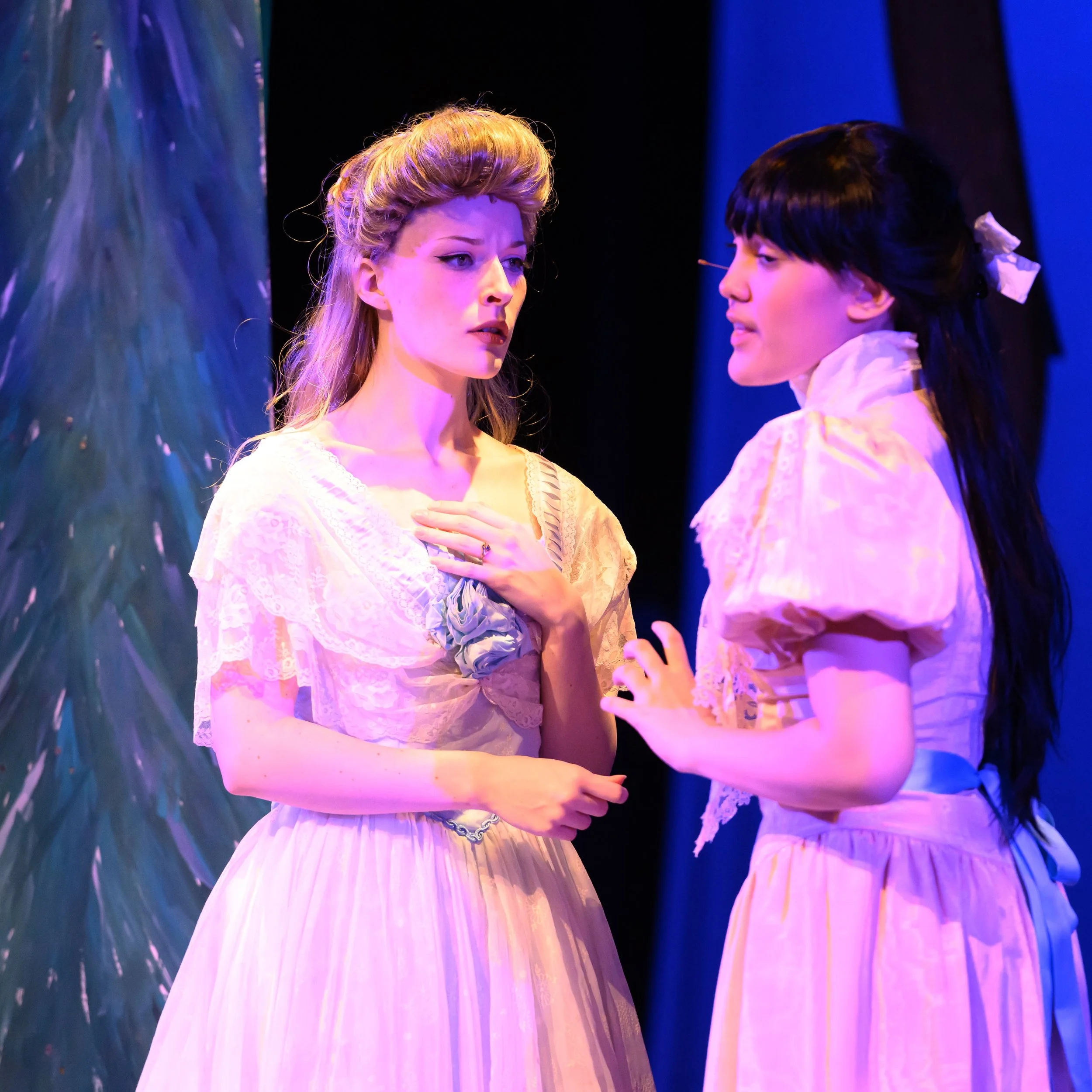 A young blonde woman in a pale blue ballgown stands listening to a black-haired teenager in a pink dress anxiously telling her something.