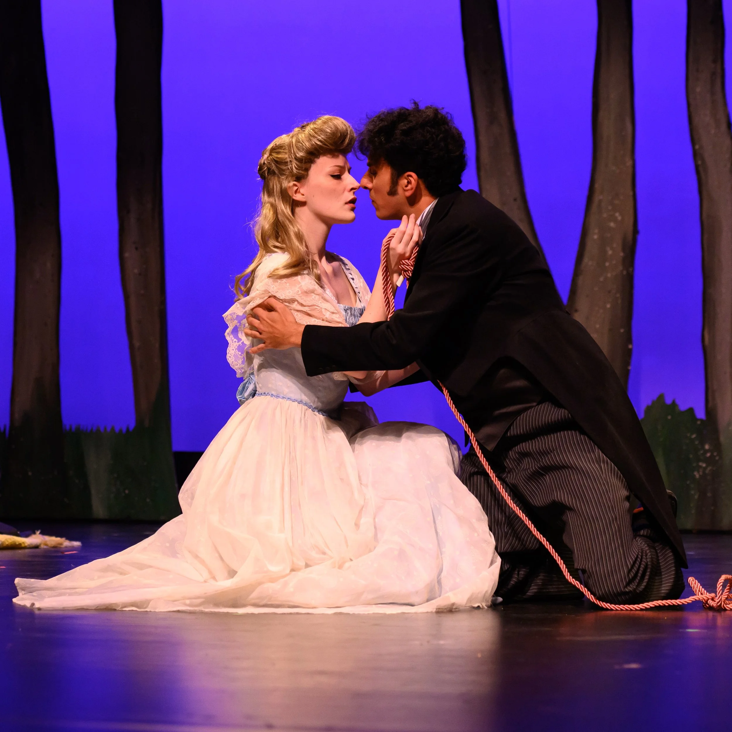 A woman in a blue ballgown and a man in black coattails kneel before each other on the ground, reaching for one another as they nearly kiss, illuminated by a blue background silhouetted with trees.