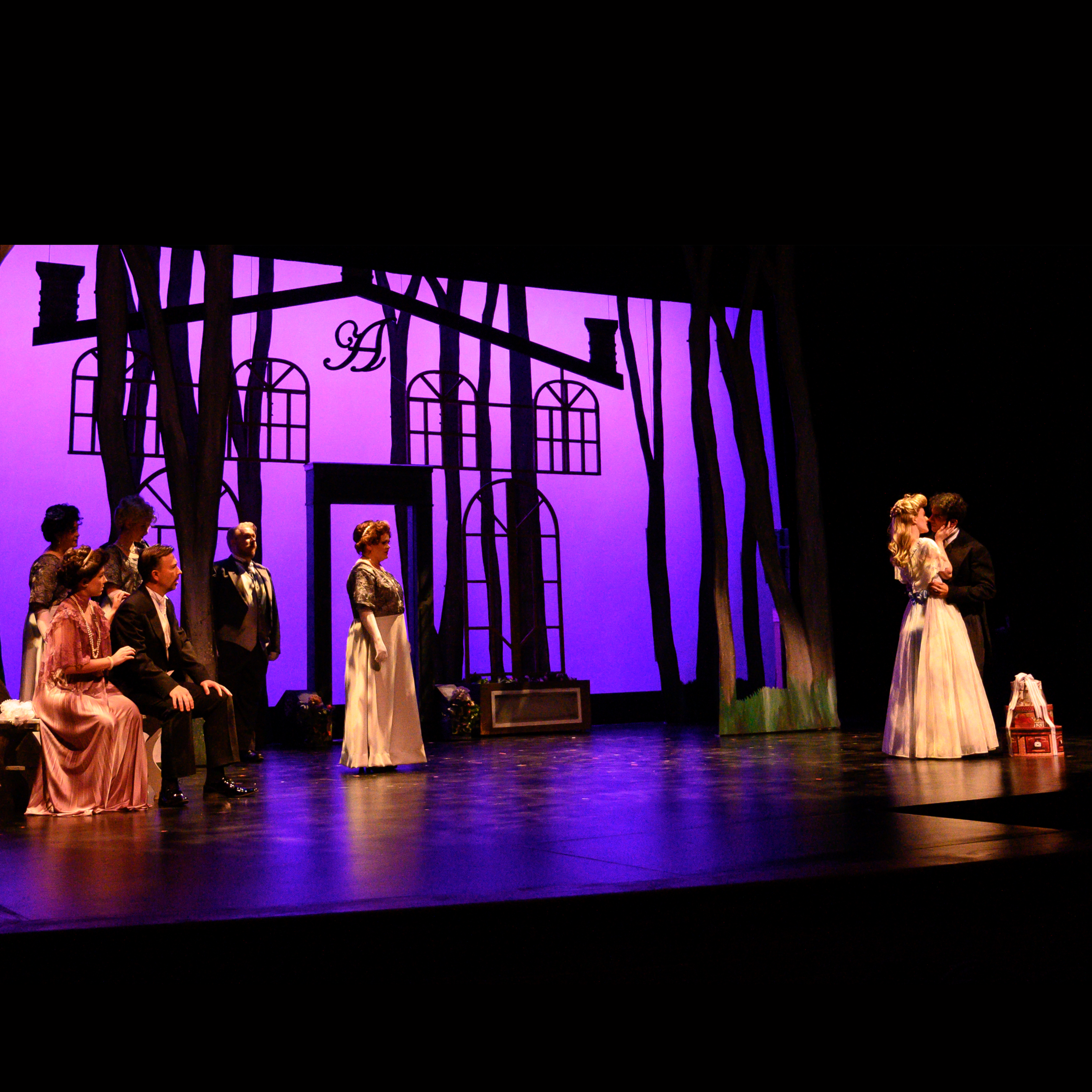 A man and woman shadowed by the quintet members, look on in surprise as a young couple kiss while they run away together, oblivious to the others on stage, illuminated by a purple background covered in the shadows of trees.