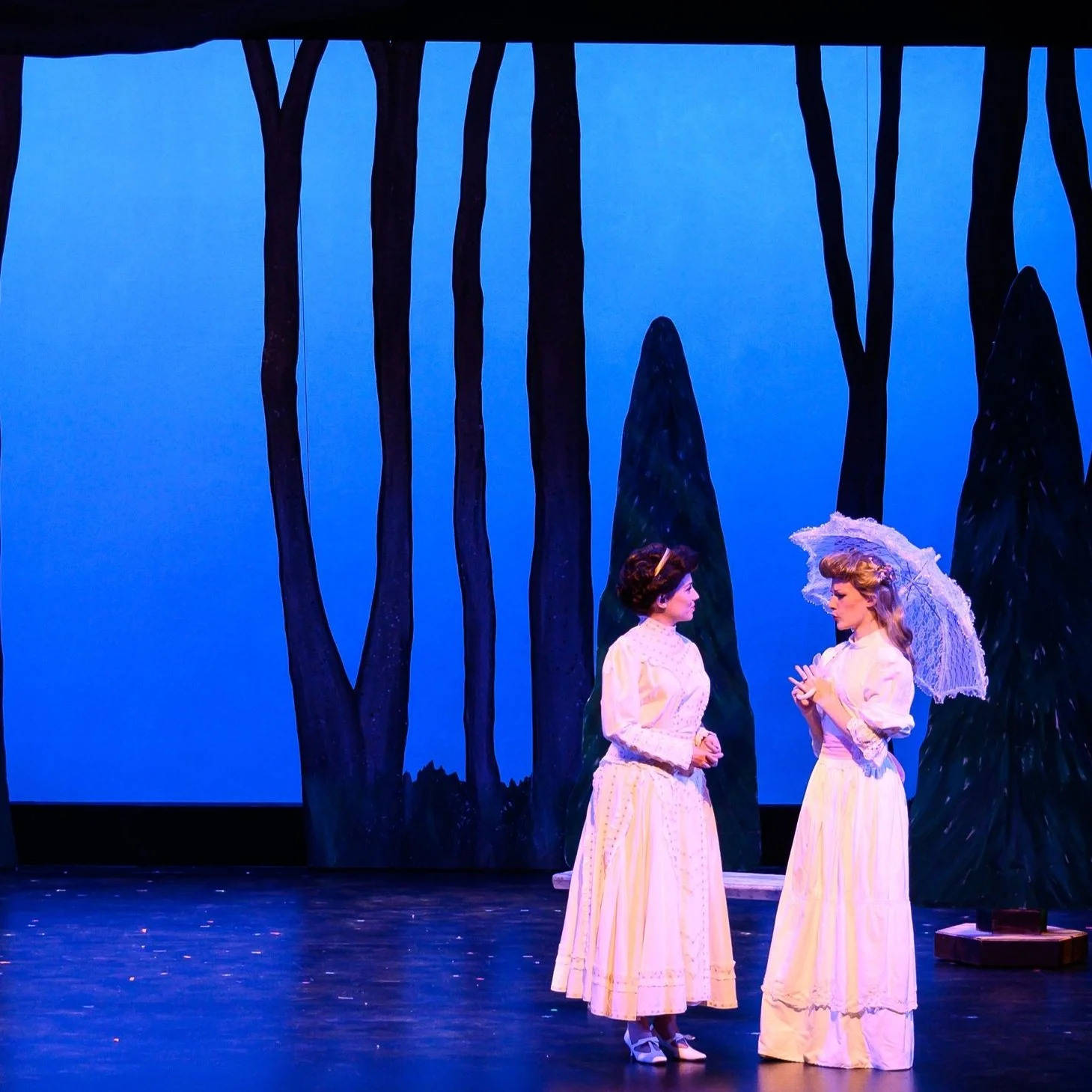 A brunette woman in a white dress wears a pinched smile next to a young blonde woman in a longer white dress holding a white lace parasol, a regretful look on her face, both standing before a blue background dotted with trees.