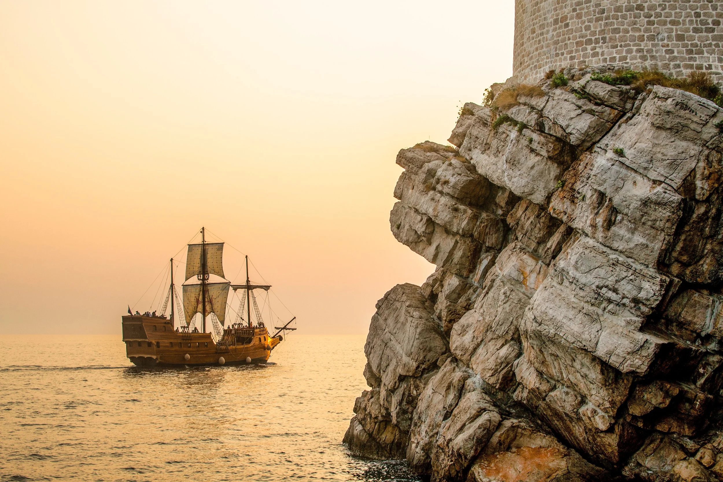 Replica pirate ship sailing around a rocky cliff, off into the sunset