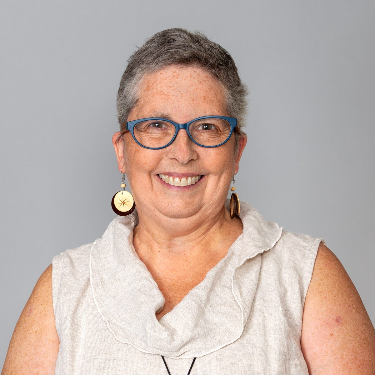 White woman in her late 50s, with short cropped grey hair in a brown linen tank.