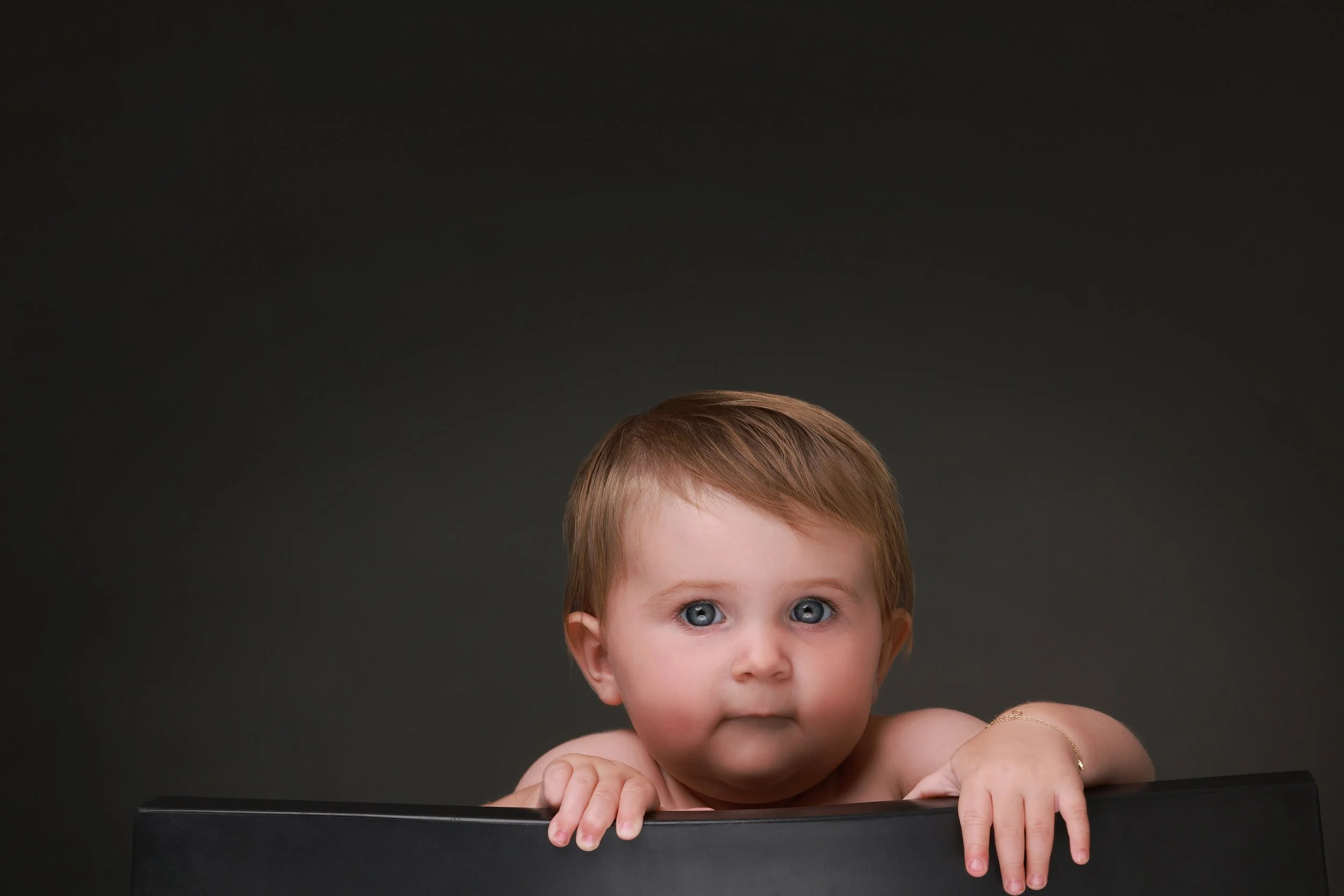 A young child with blue eyes and light brown hair looking over the edge of a black surface against a dark gray background.