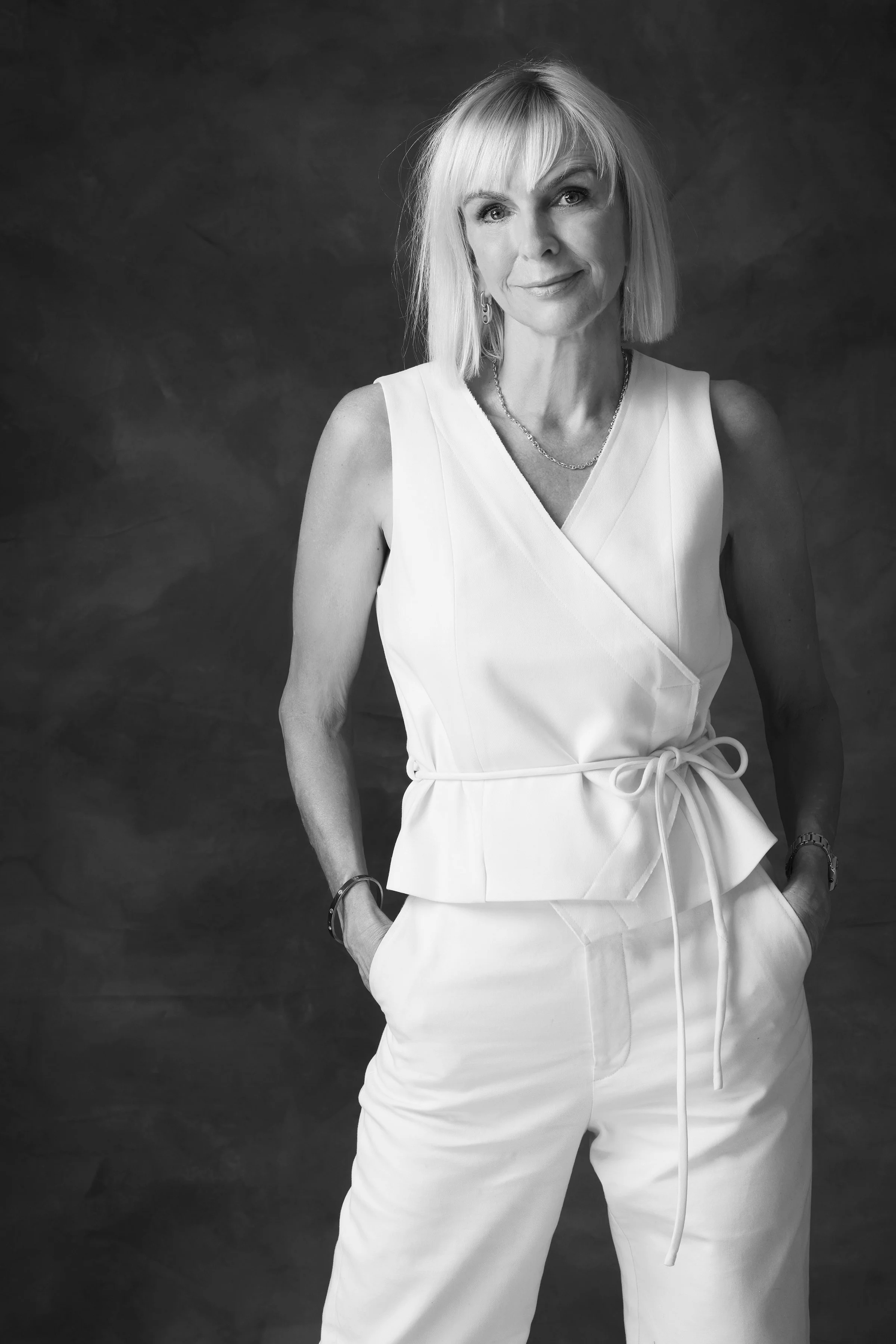 Black and white portrait of a mature woman with blond hair, wearing a sleeveless white top and matching pants, standing with her hands in her pockets against a dark background.