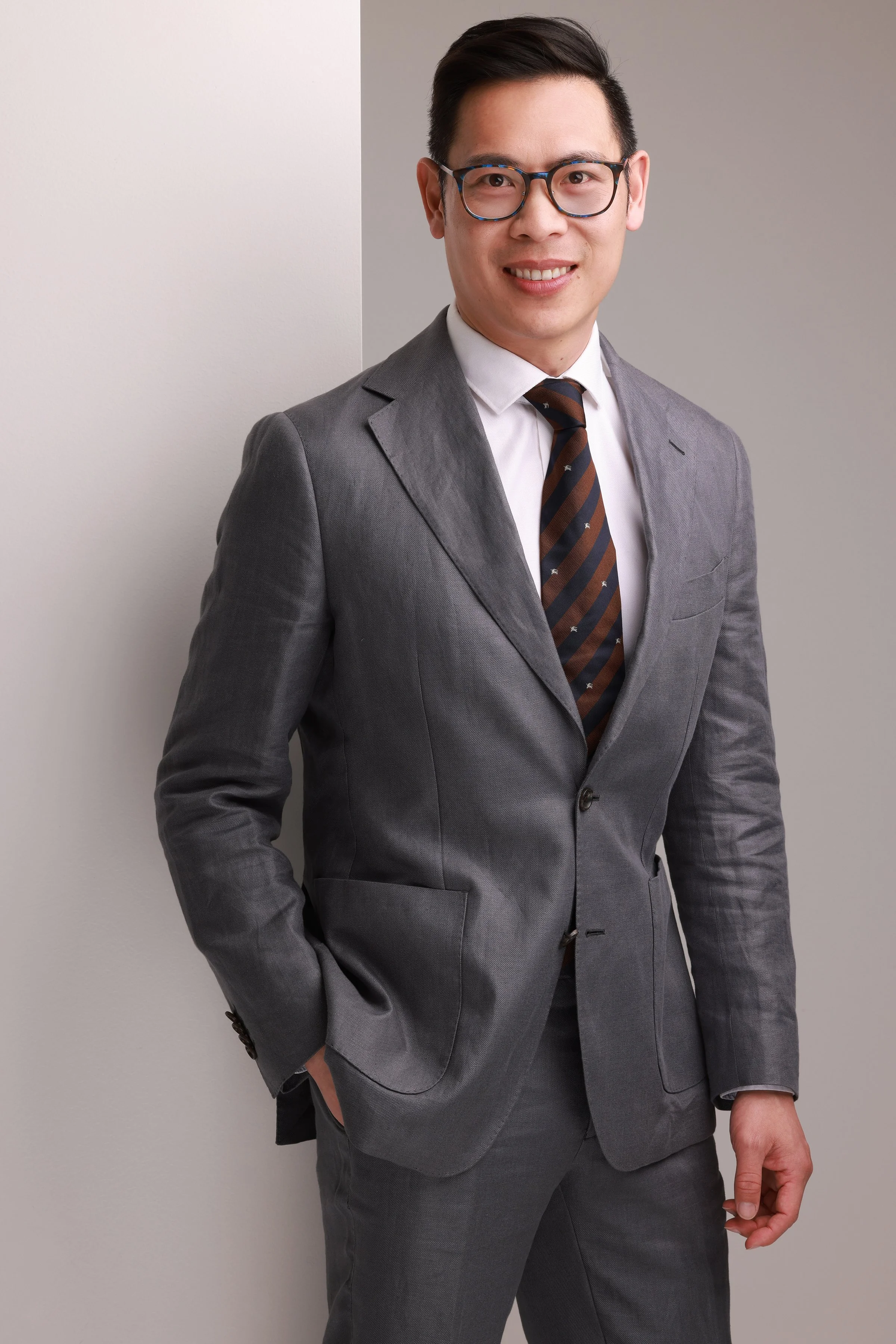 A man in a gray suit, white shirt, and brown striped tie standing against a plain background with a smile.