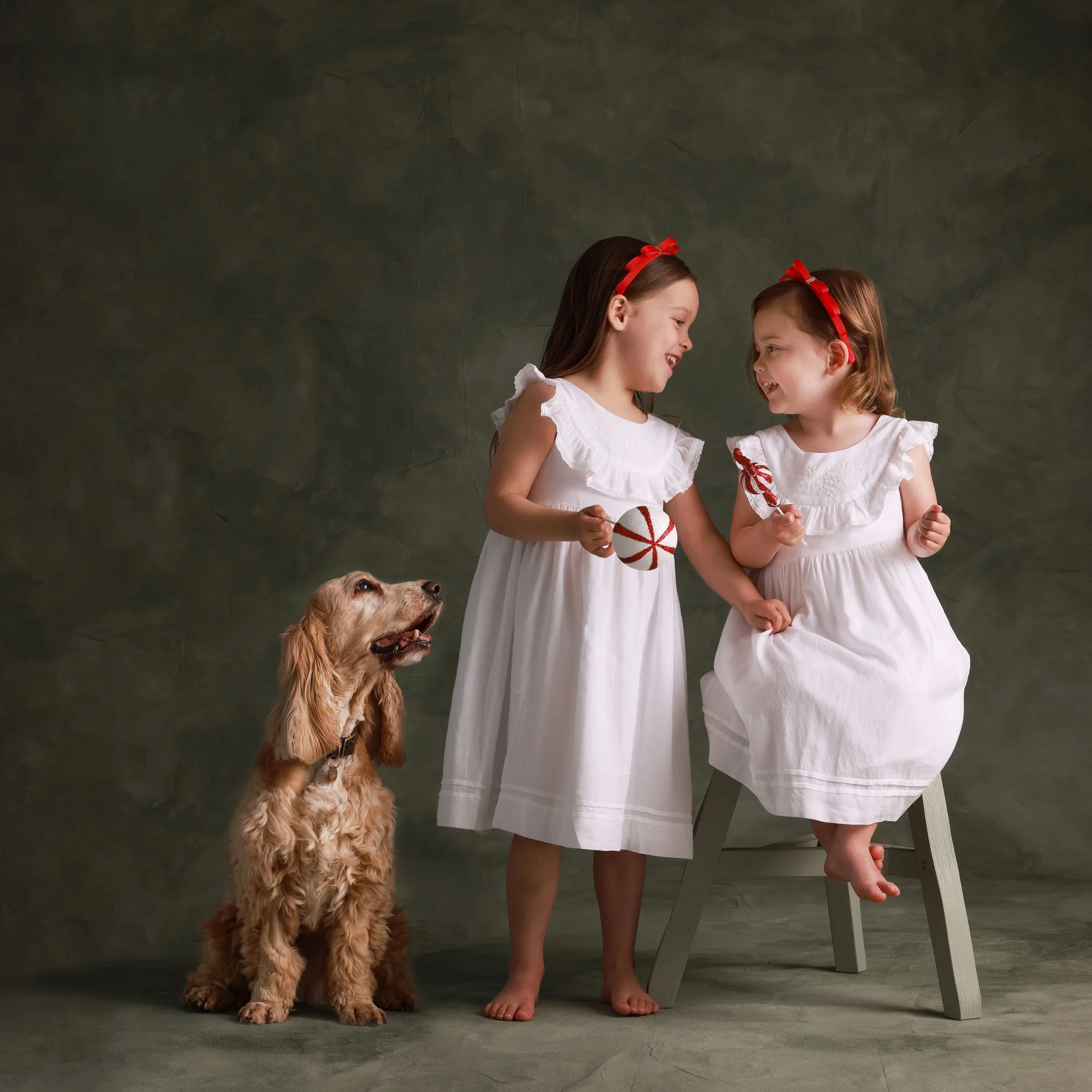 Two young girls in white dresses with red headbands smiling and holding Christmas ornaments, with a golden cocker spaniel sitting nearby on a dark gray background.