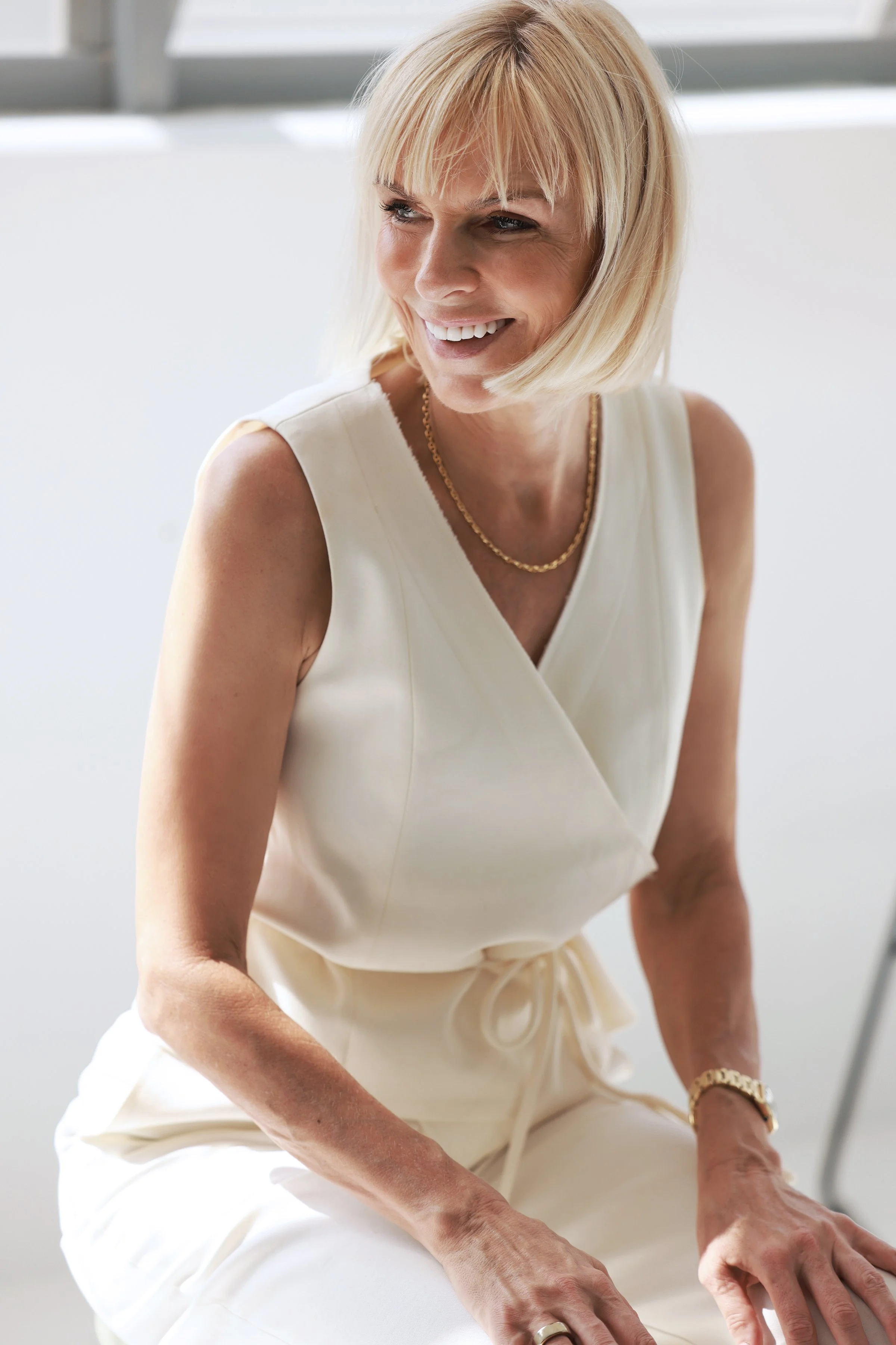 A smiling blonde woman in a sleeveless white dress with a gold necklace, sitting in a bright room with large windows.