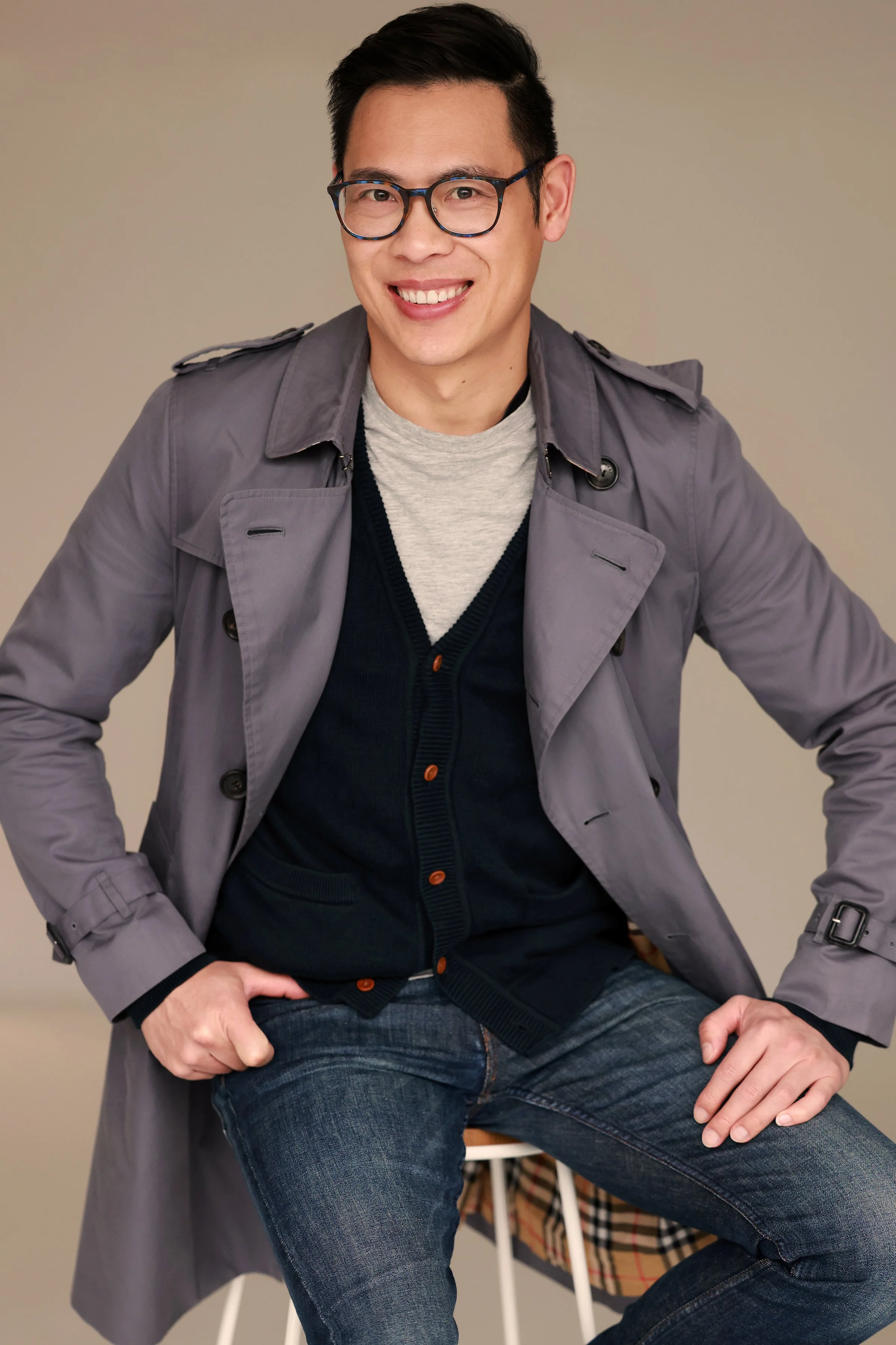 A man smiling, wearing glasses, a gray trench coat, a navy cardigan, a light gray sweatshirt, and jeans, sitting on a bench against a plain beige background.