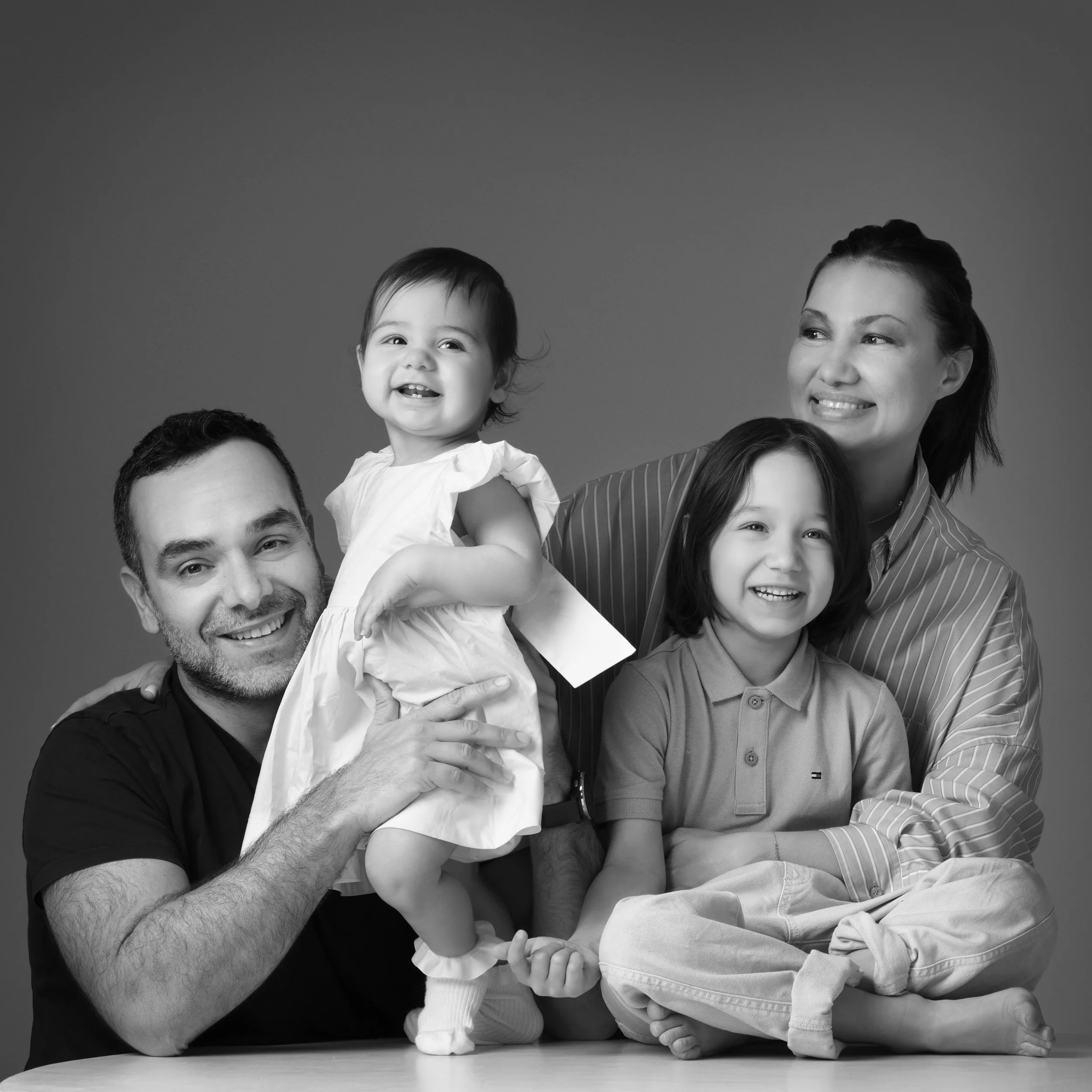 A family of four smiling poses in a studio, with a man holding a young girl on his shoulder, a woman sitting beside a boy, all in black and white.