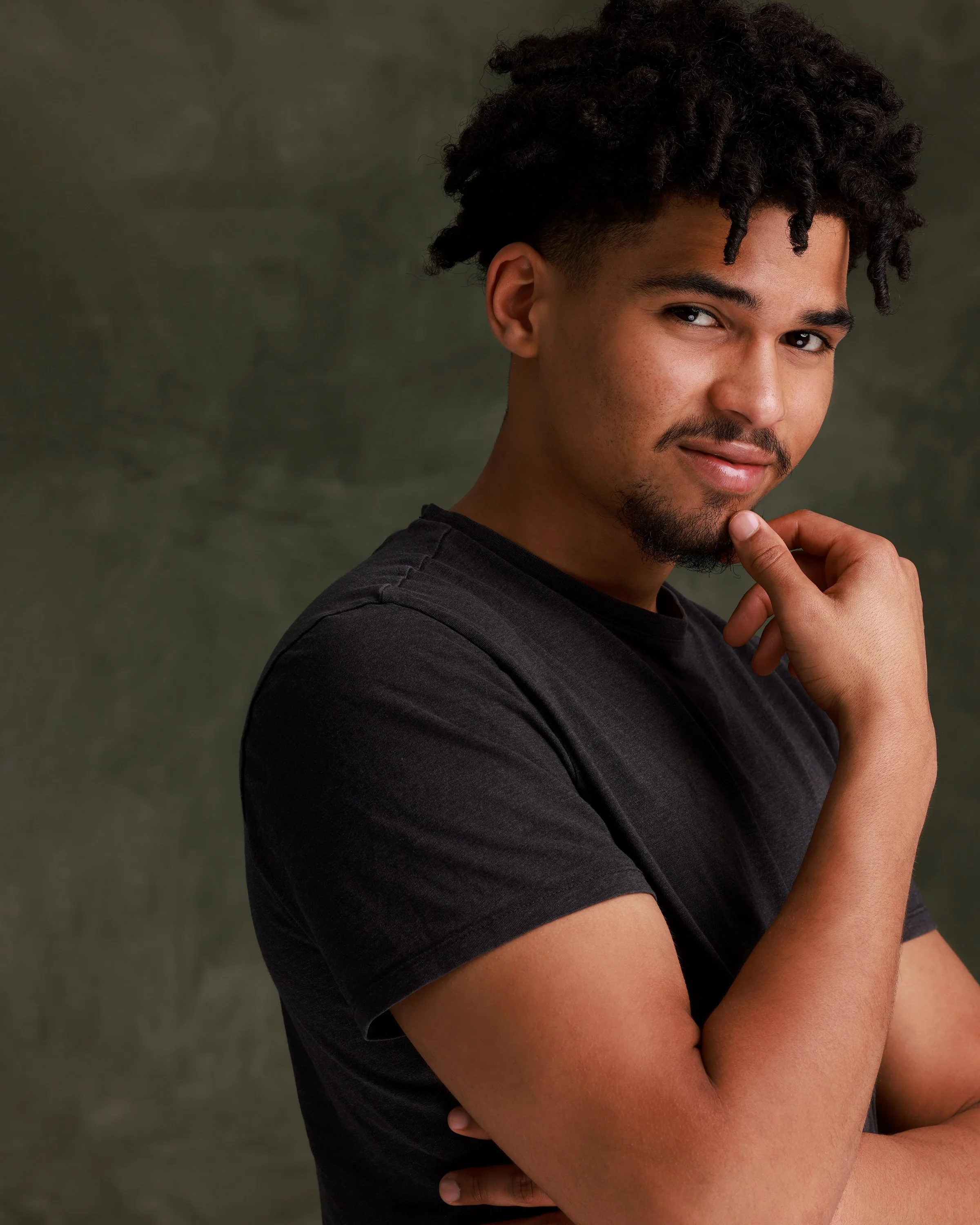 A young man with curly hair, a goatee, and a black t-shirt, posing thoughtfully with one hand on his chin against a muted gray-green background.