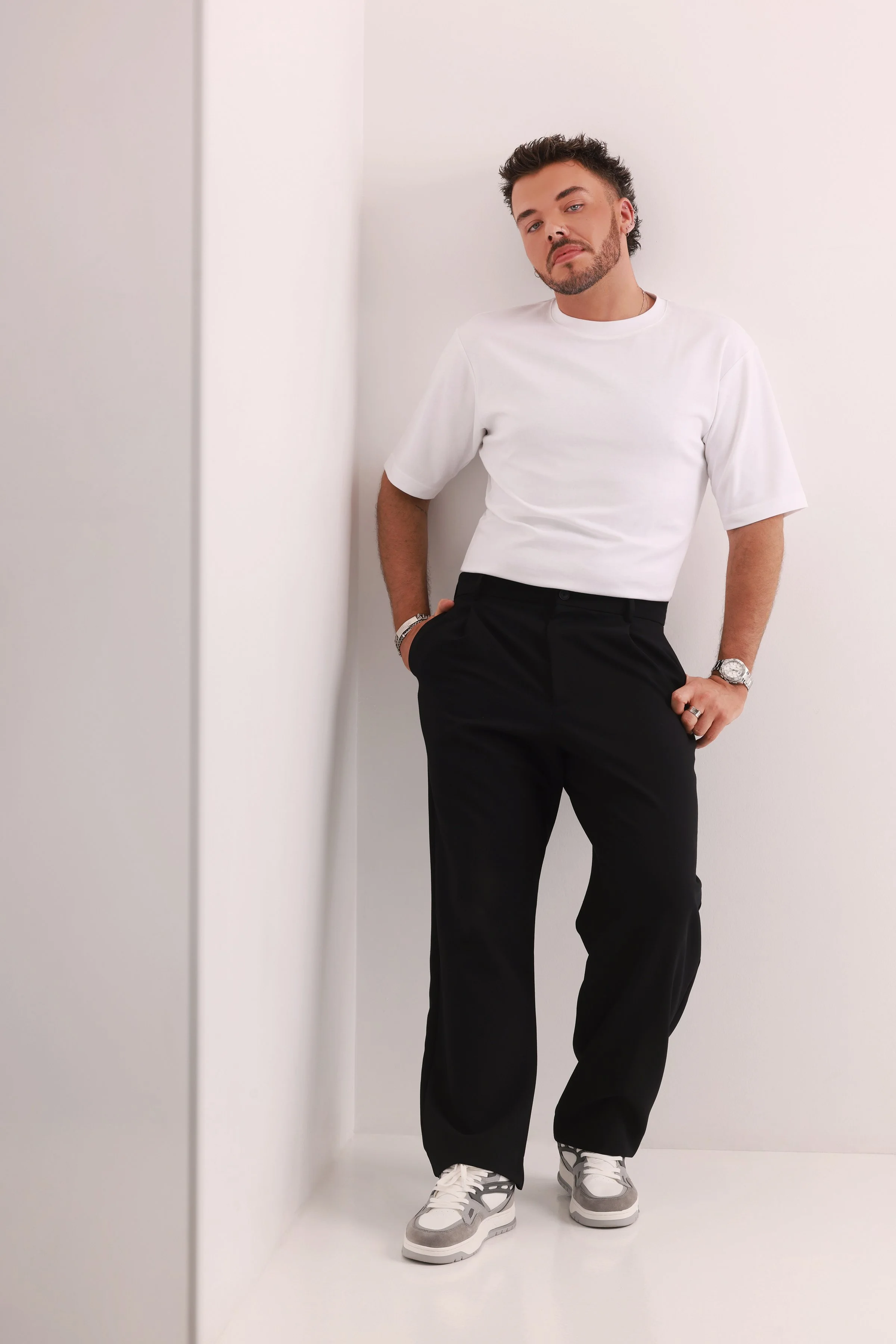 A man with a beard and styled hair stands casually in a corner, wearing a white t-shirt, black pants, and white sneakers, against a plain white background.