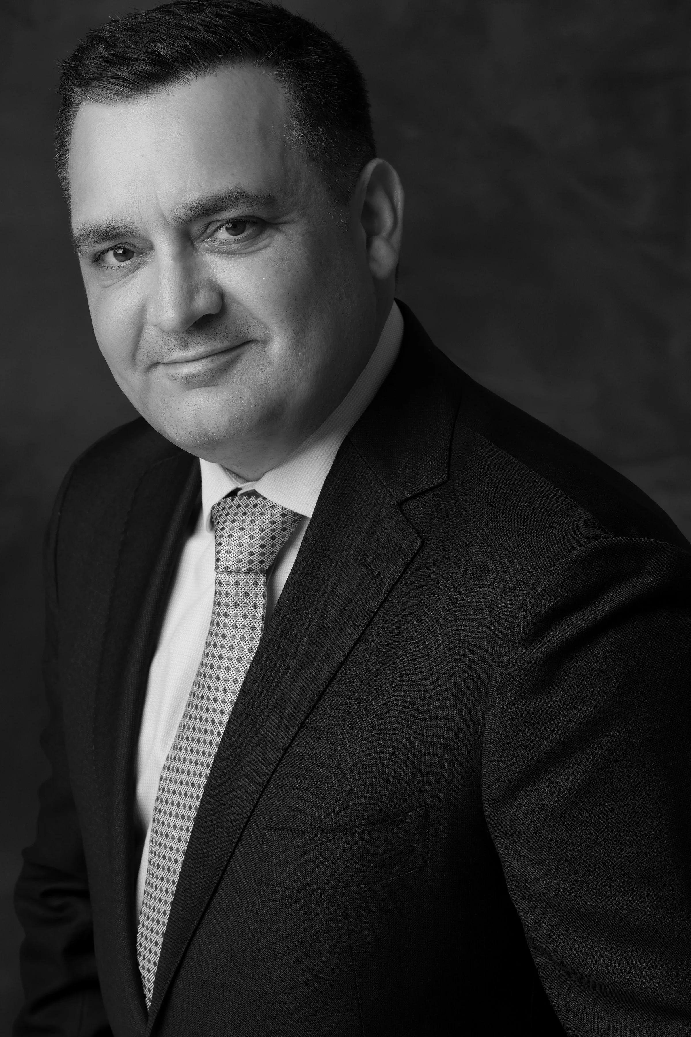 Black and white professional portrait of a man in a suit and tie, smiling slightly, with a dark background.