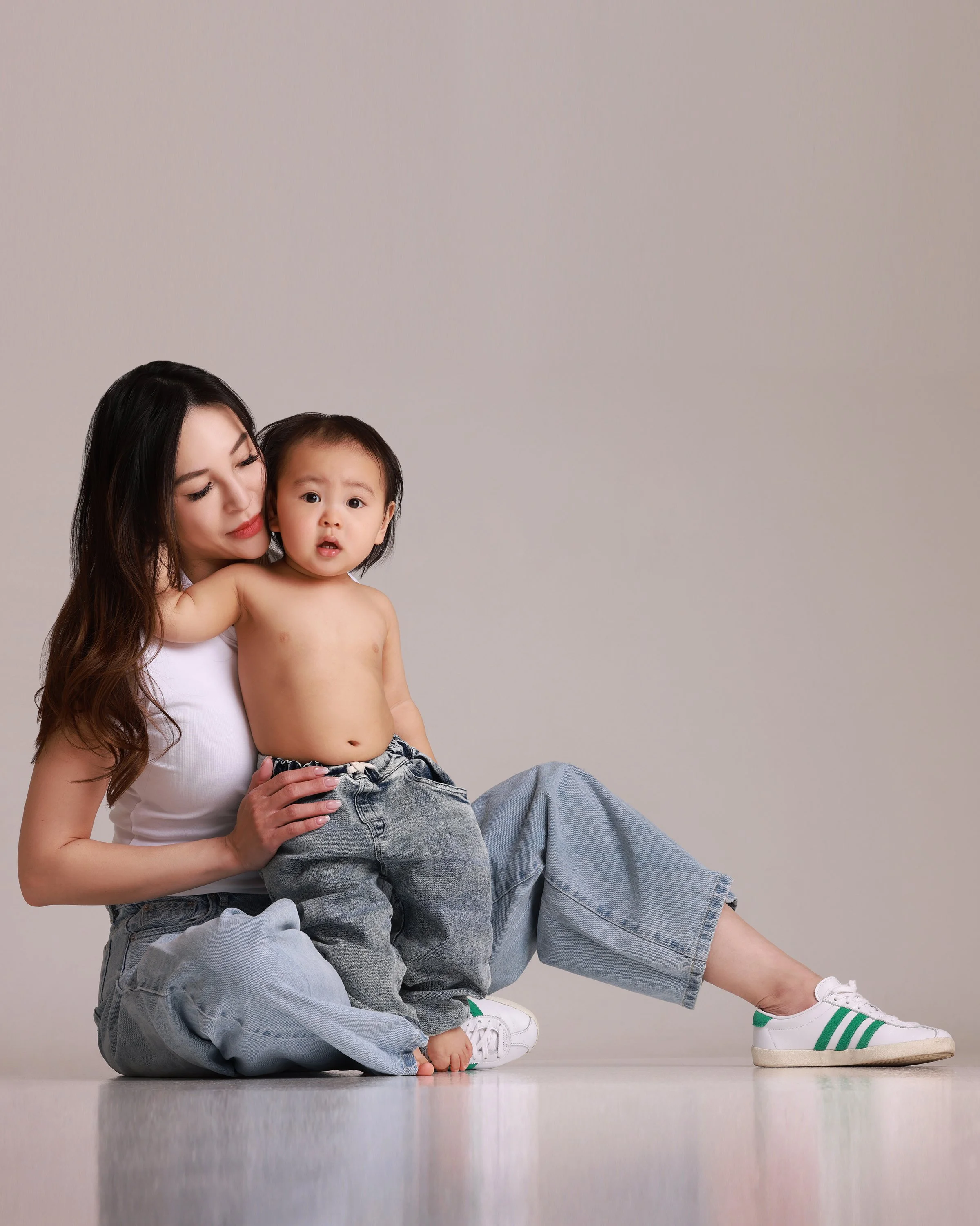 A woman with long dark hair sitting on the floor with a shirtless young boy in her lap. She wears a white tank top, baggy blue jeans, and white sneakers with green stripes. The boy appears surprised and is wearing loose gray jeans, and is barefoot. T