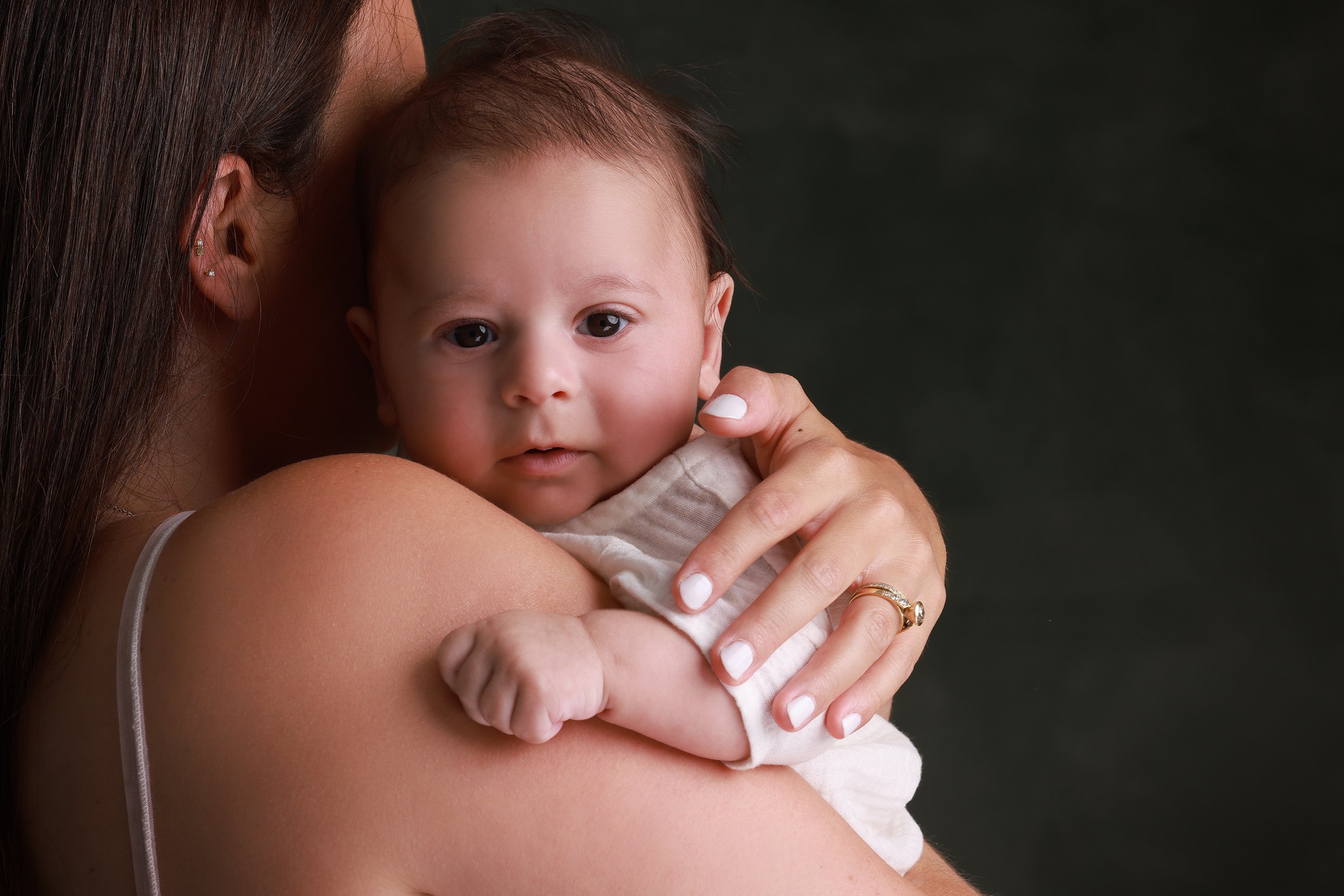 Close-up of a mother holding her young baby against her shoulder, with the baby's face visible and arms wrapped around her. The mother has dark hair and multiple earrings, and is wearing a gold ring.