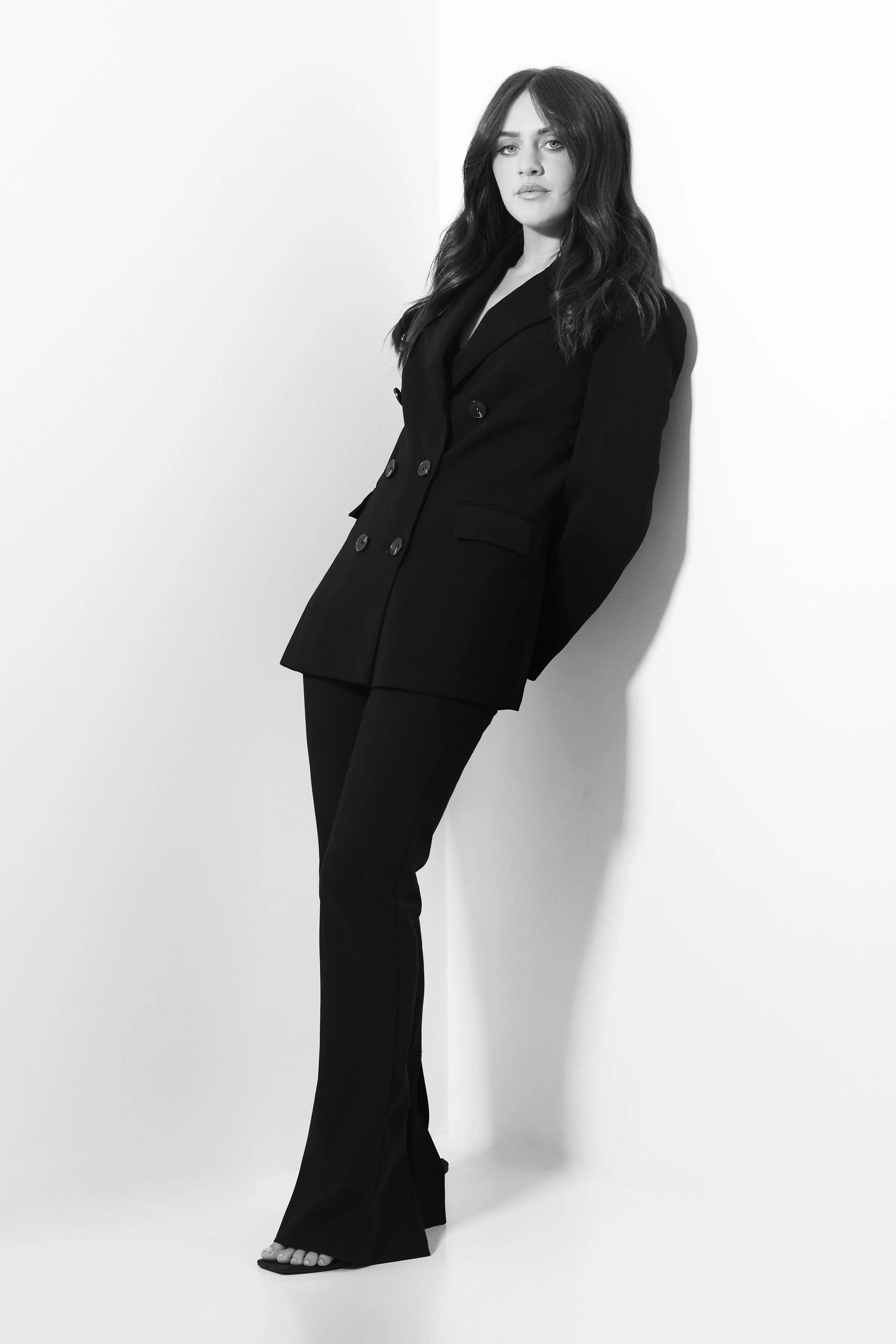 A woman with long wavy hair wearing a black blazer and pants, standing against a white background.