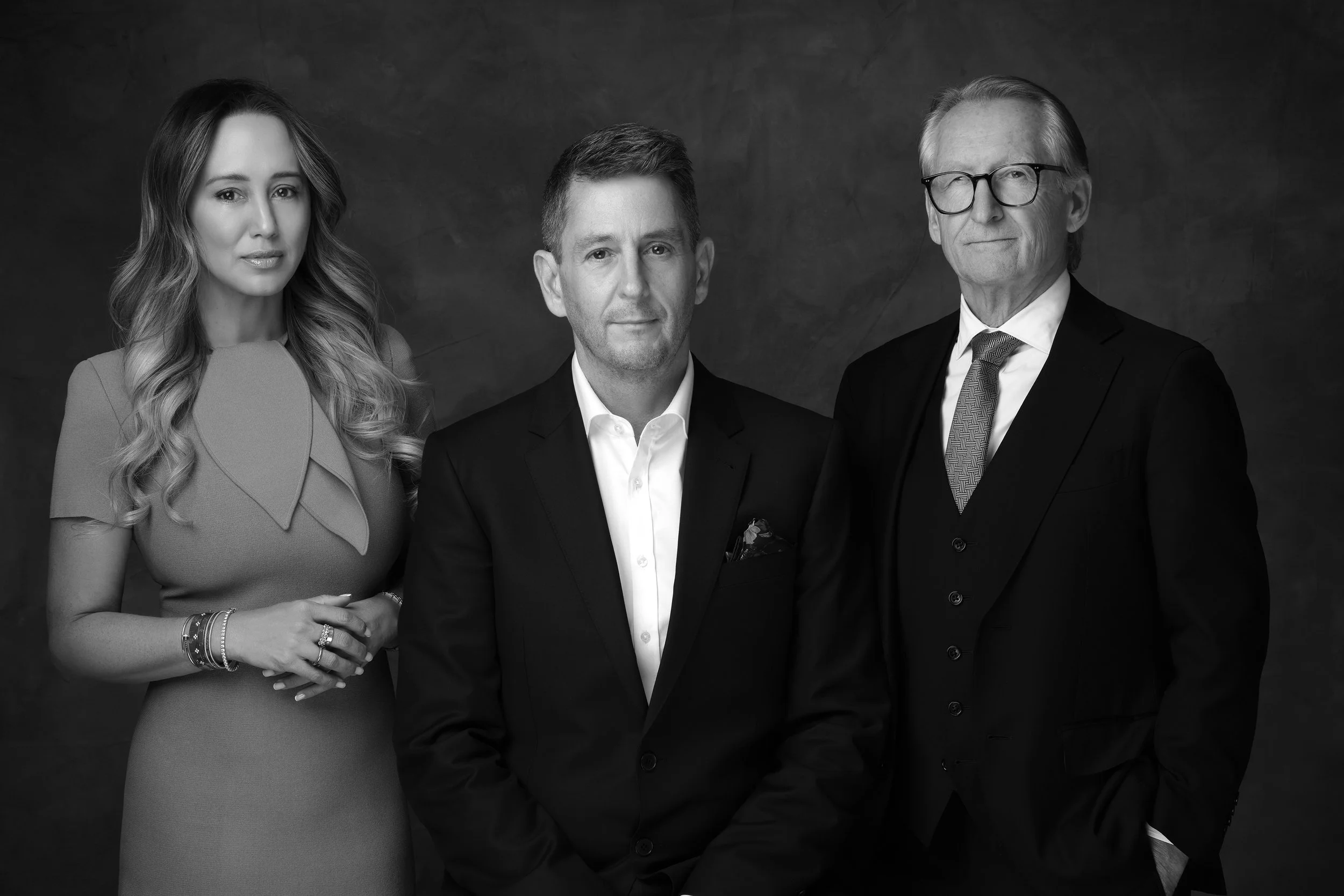 Black and white portrait of three professionally dressed individuals, two men and one woman, standing against a dark background.