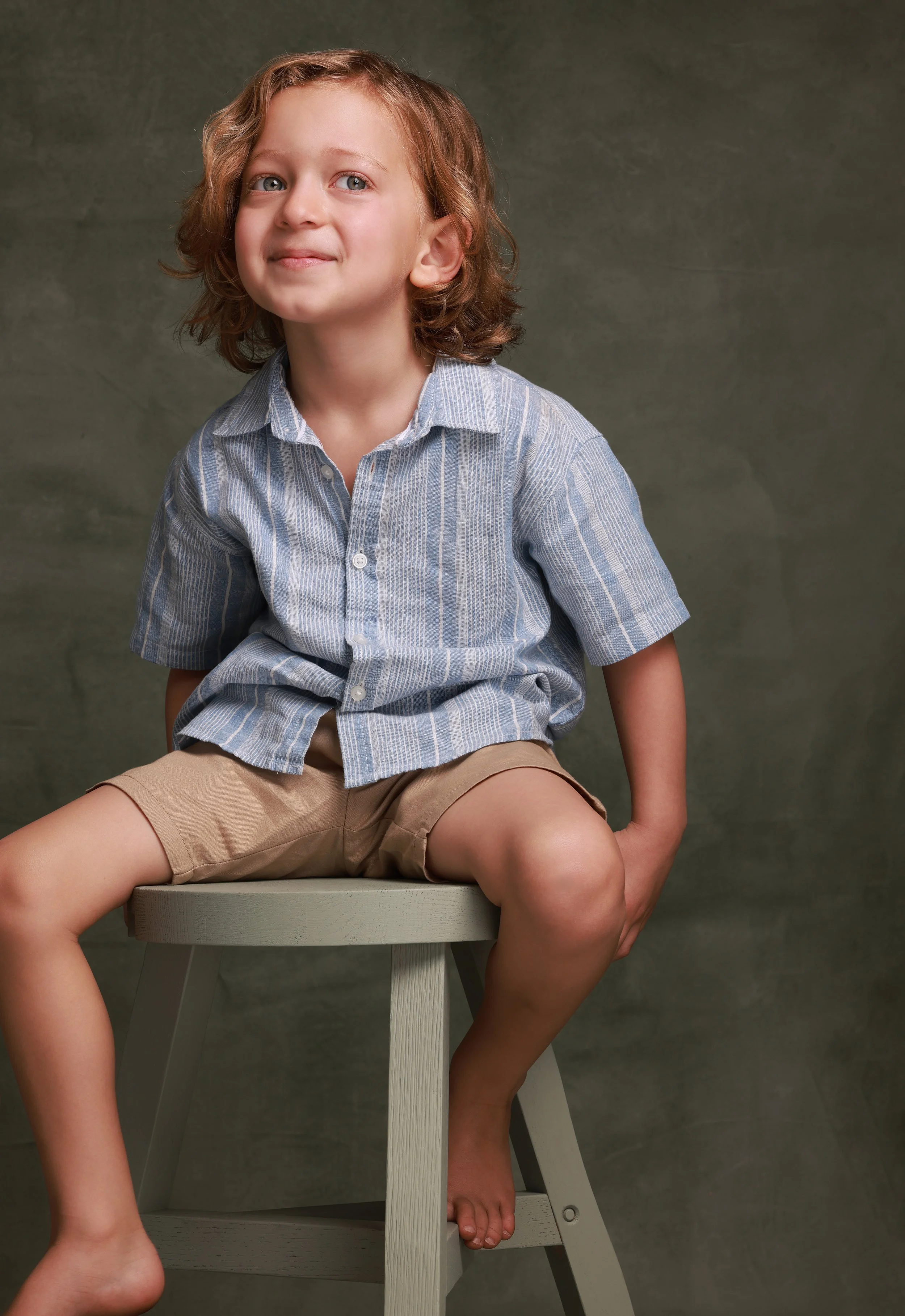 A young boy with curly, light brown hair and blue eyes sitting barefoot on a stool against a dark background, wearing a blue and white striped short-sleeve button-up shirt and beige shorts.