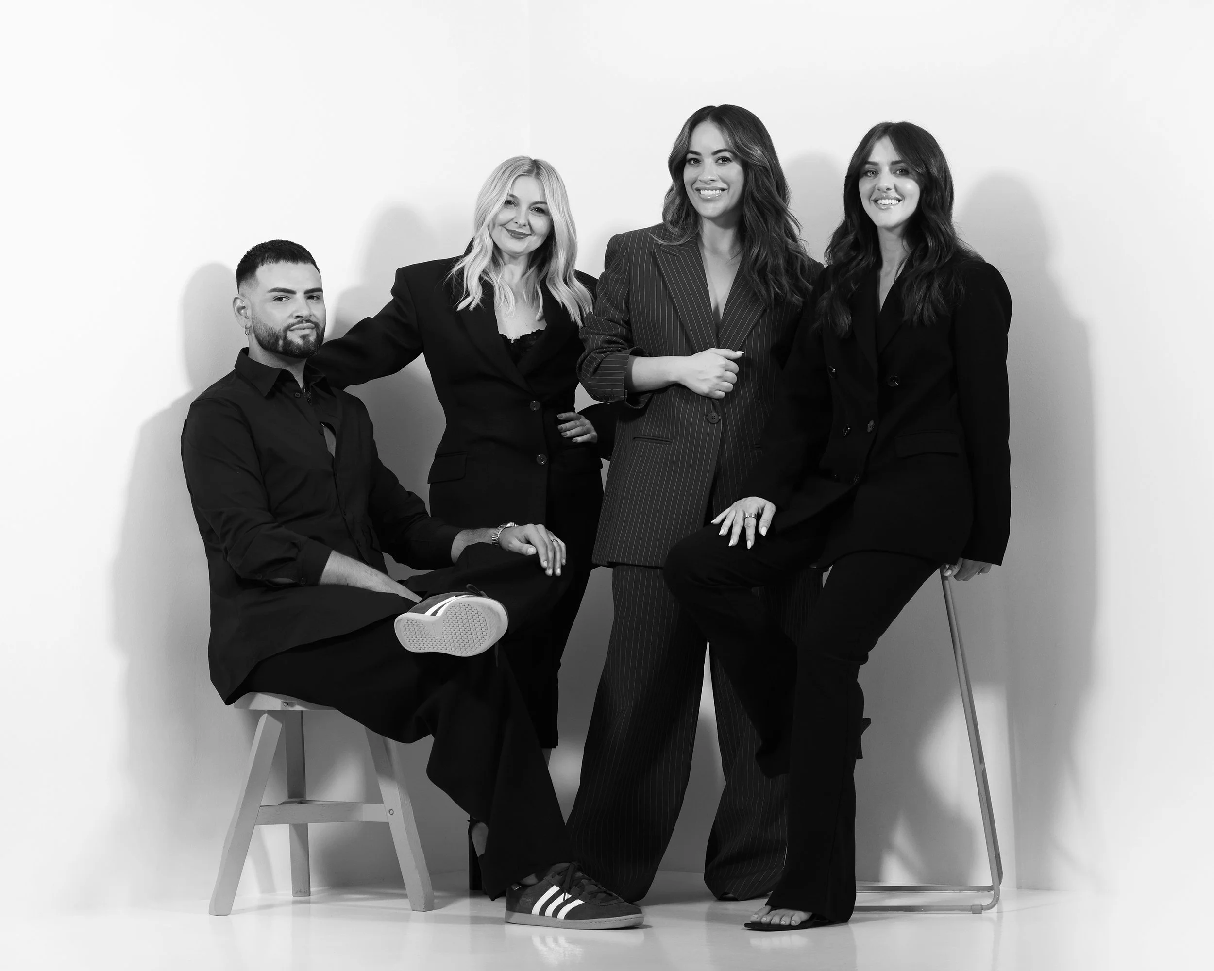 Group of four diverse professionals in black outfits, posed against a white background, with two sitting on chairs and two standing, smiling for the camera.