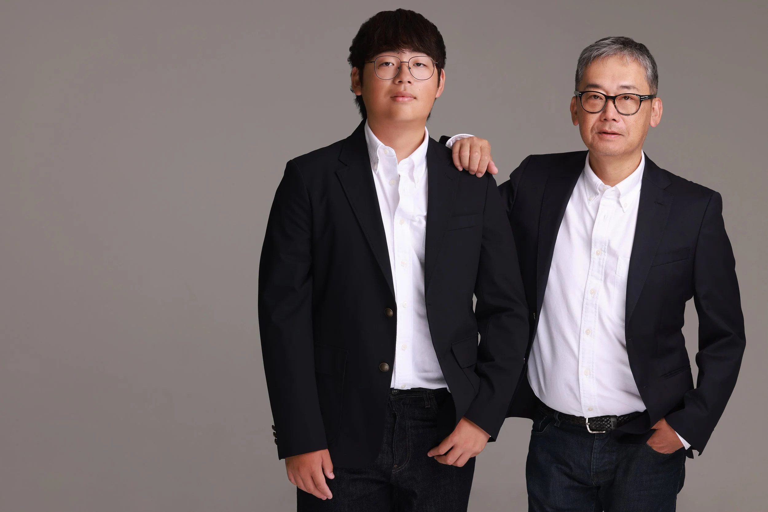 Two men in black suits and white shirts standing together against a gray background, with one man's hand on the other's shoulder.