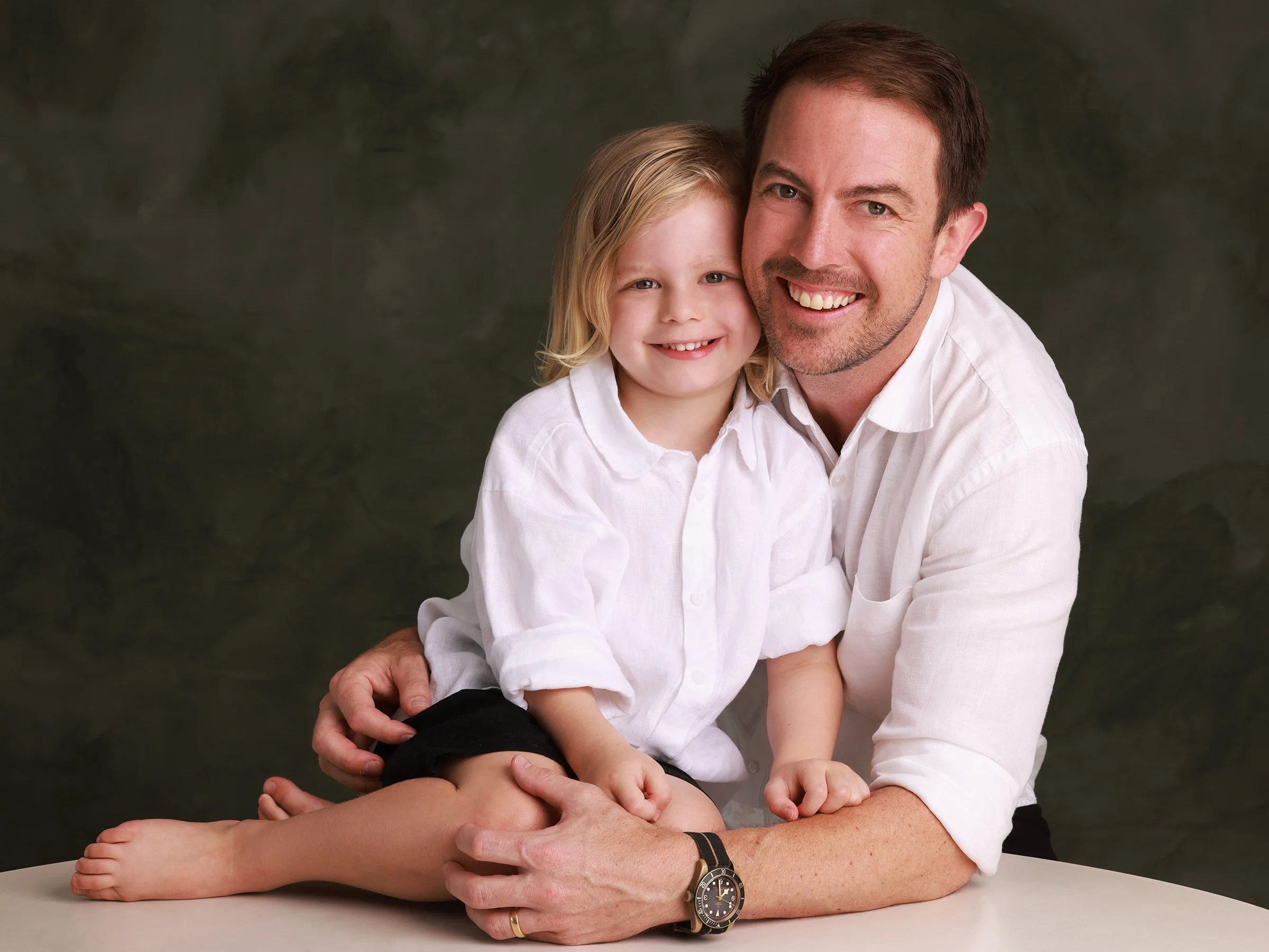 A man and a young girl sitting at a table, smiling and hugging against a dark background