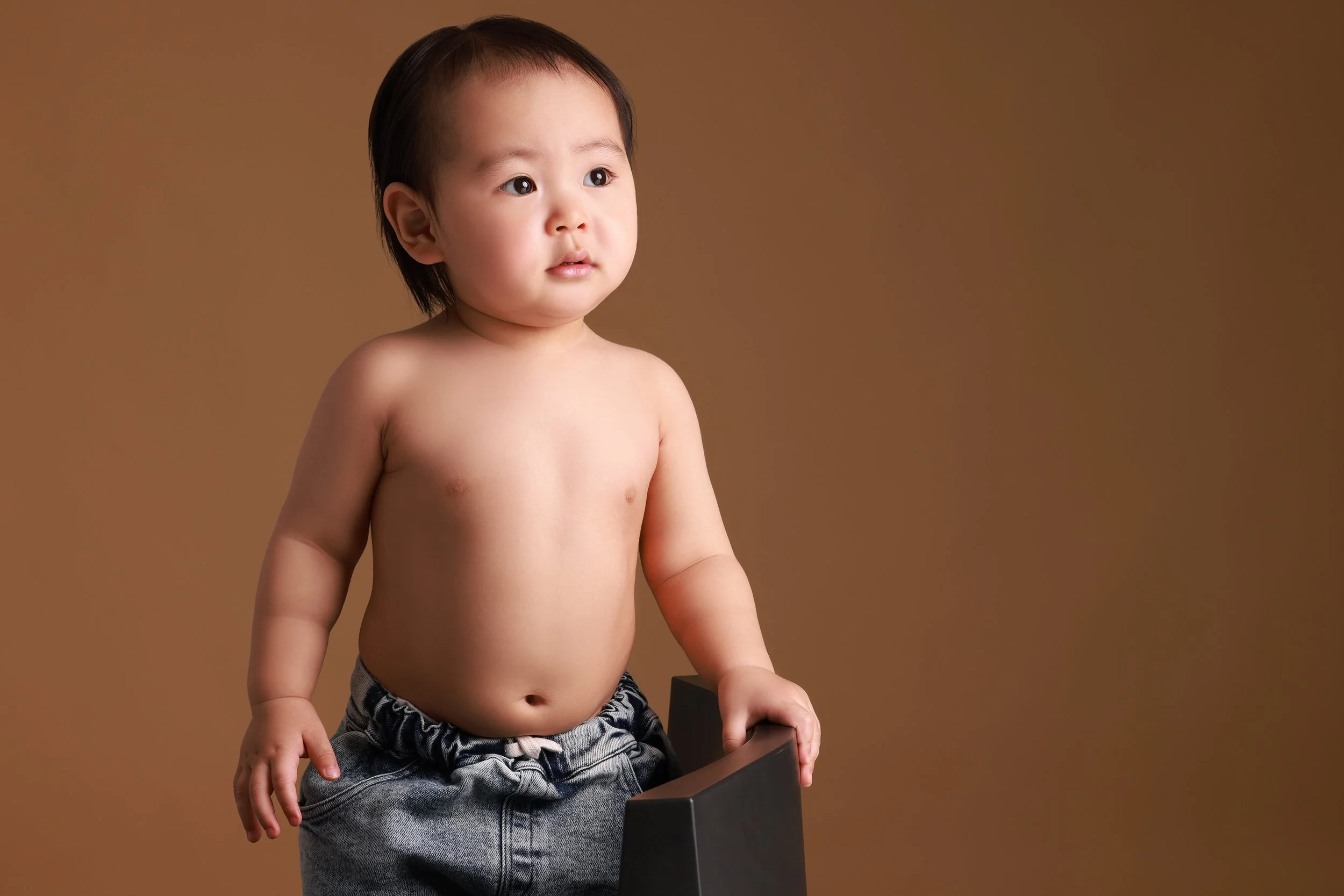 A young Asian toddler boy standing shirtless against a plain brown background, wearing grey denim shorts and holding a black object with one hand.