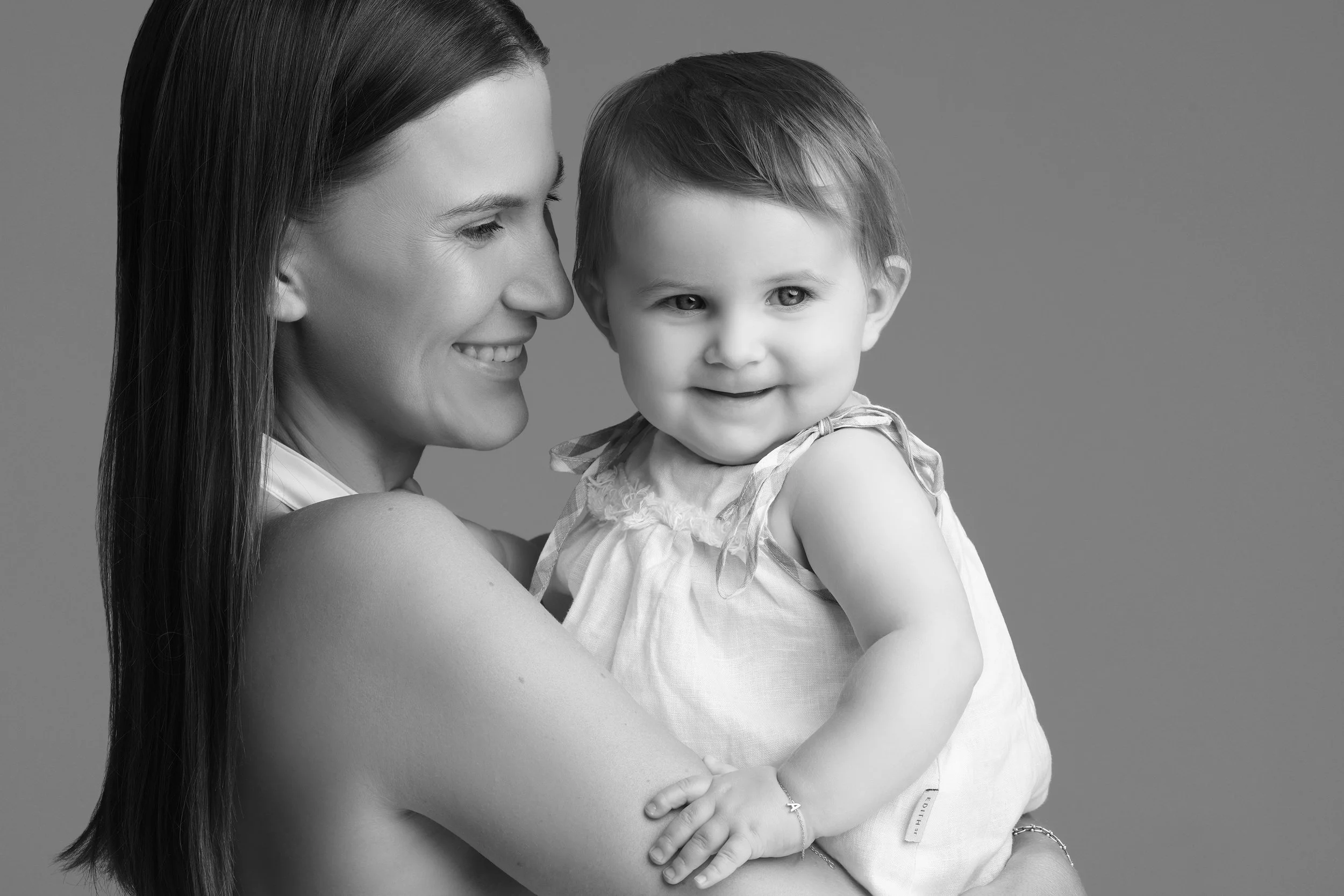 A woman holding a smiling young girl, both looking to the side, in a studio shot in black and white.