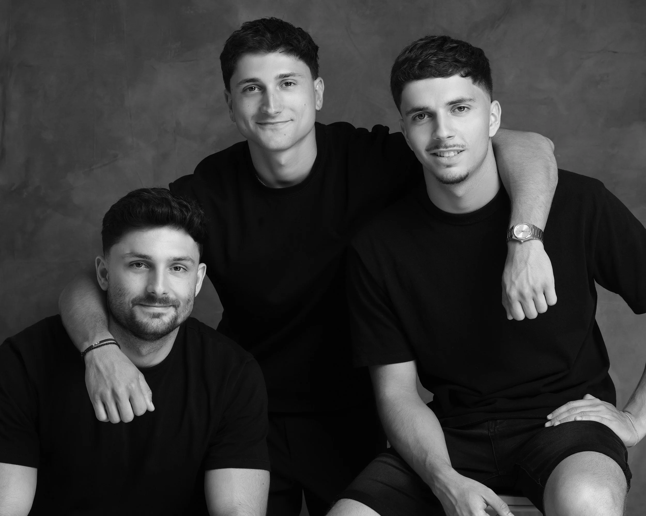 Black and white photo of three young men, two sitting and one standing behind them, with arm around each other's shoulders, smiling at the camera against a plain background.