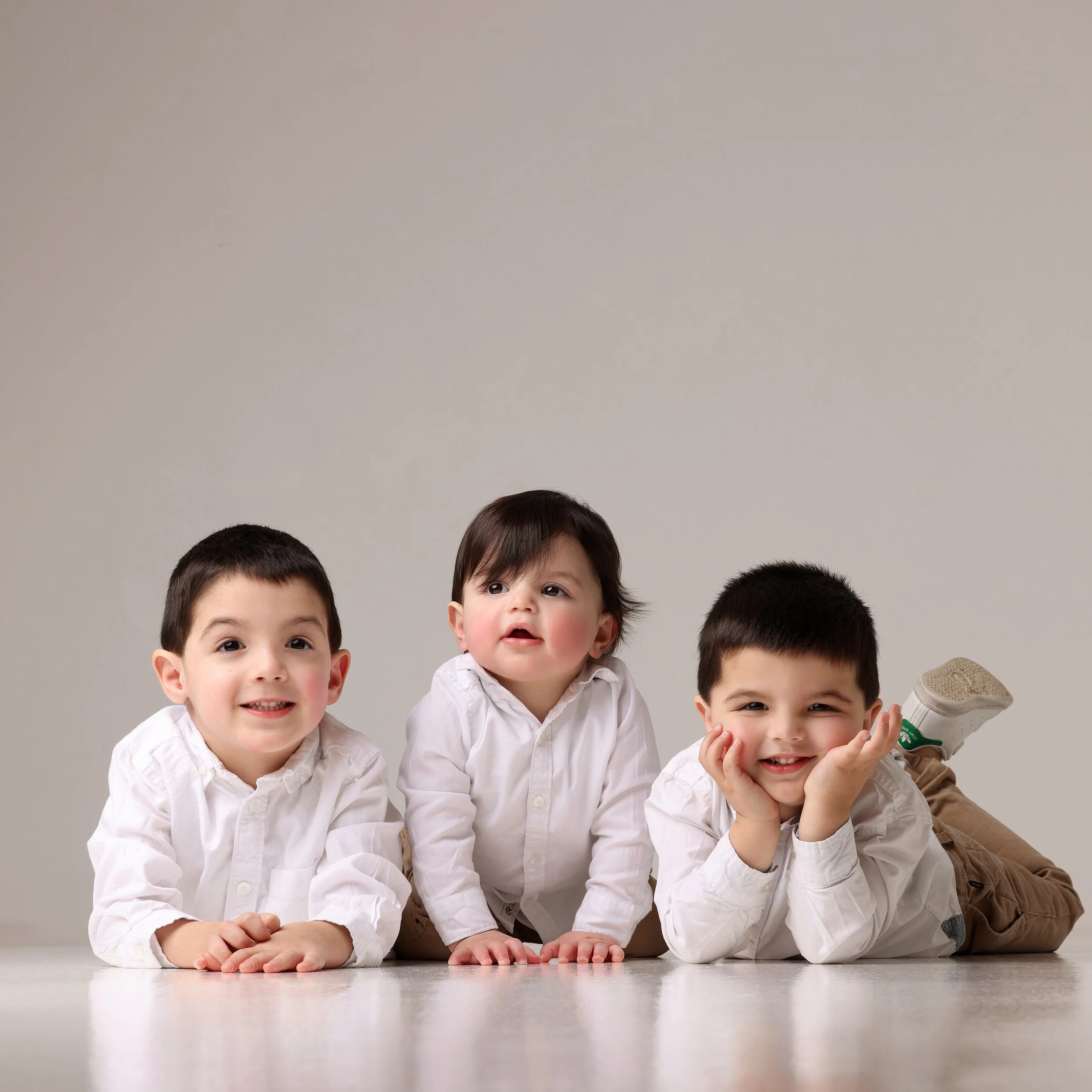 Three young boys lying on the floor, wearing white shirts, posing for a photo against a plain background.