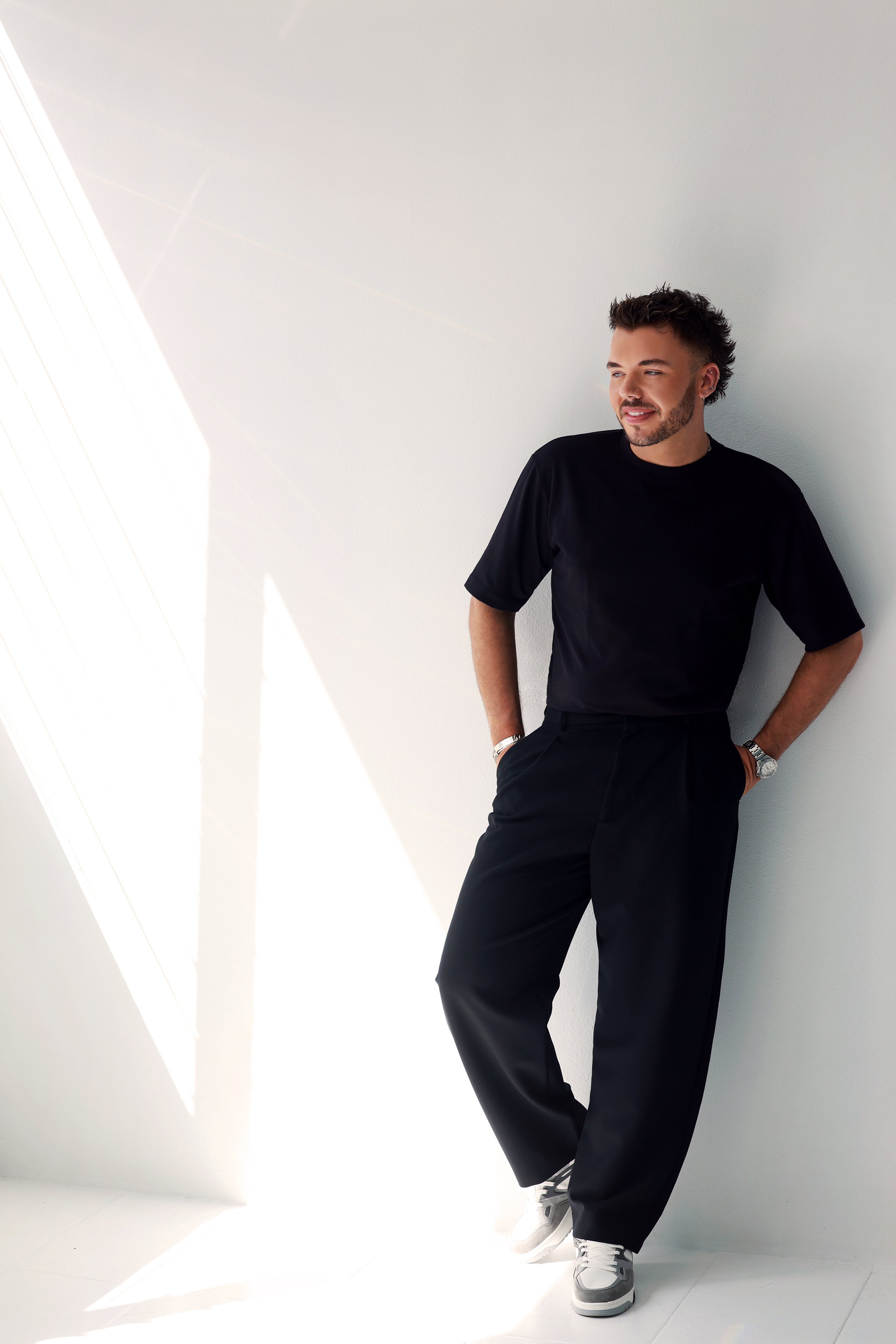 A man in a black T-shirt and black pants leaning against a white wall with sunlight creating shadows.