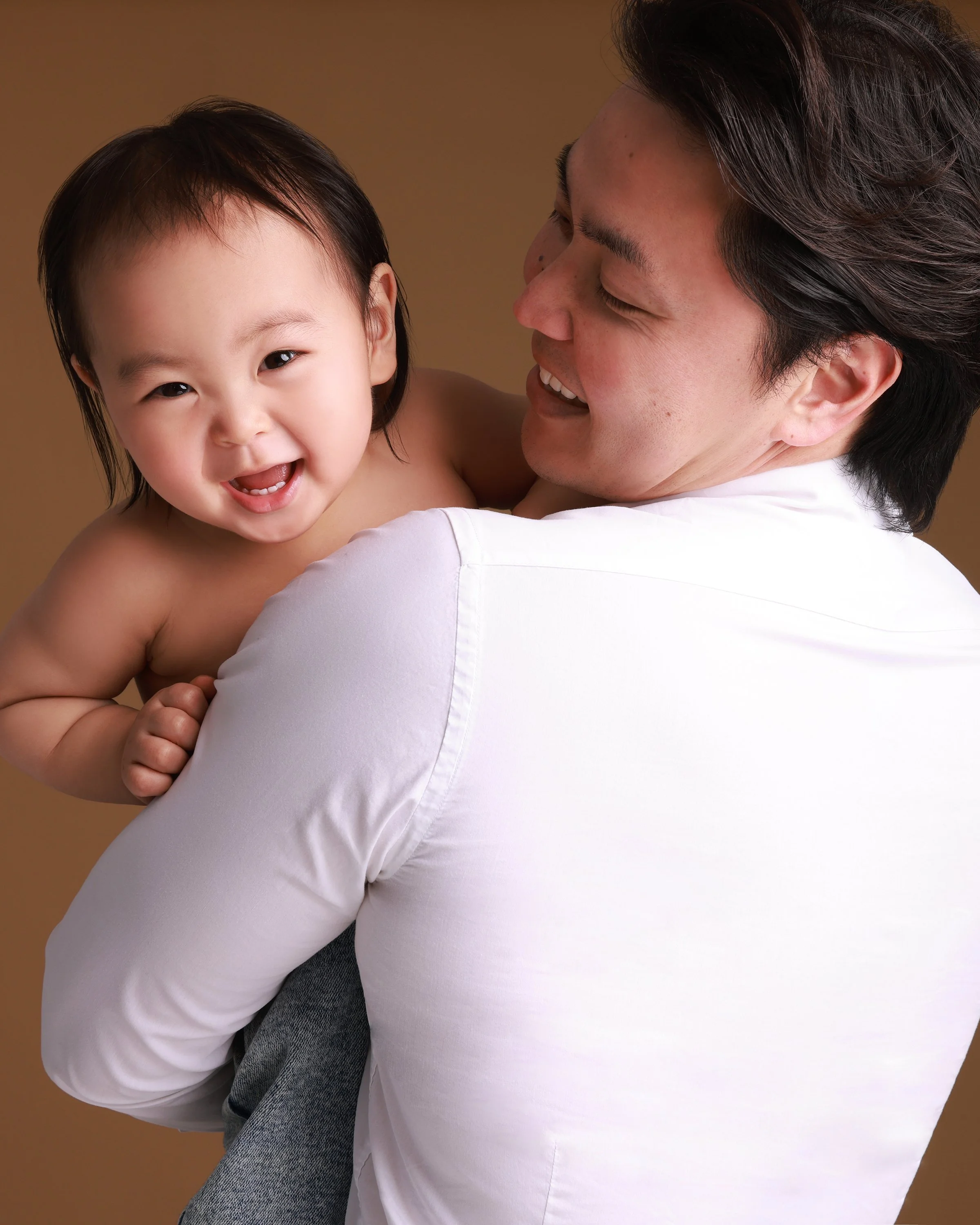 A father holding his young child and smiling at the camera against a warm background.