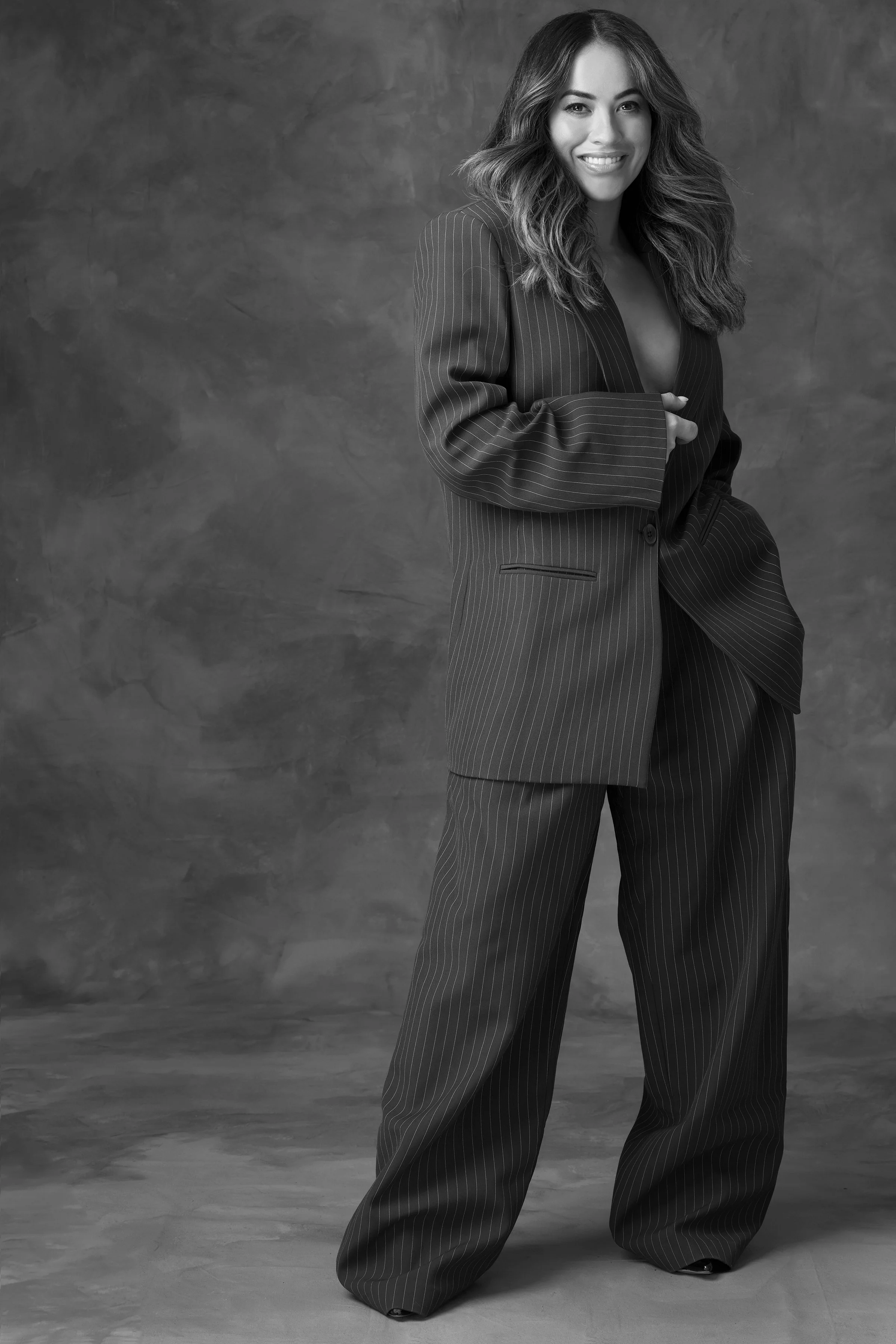 A woman with long wavy hair wearing a pinstripe suit, smiling, standing against a textured gray background.